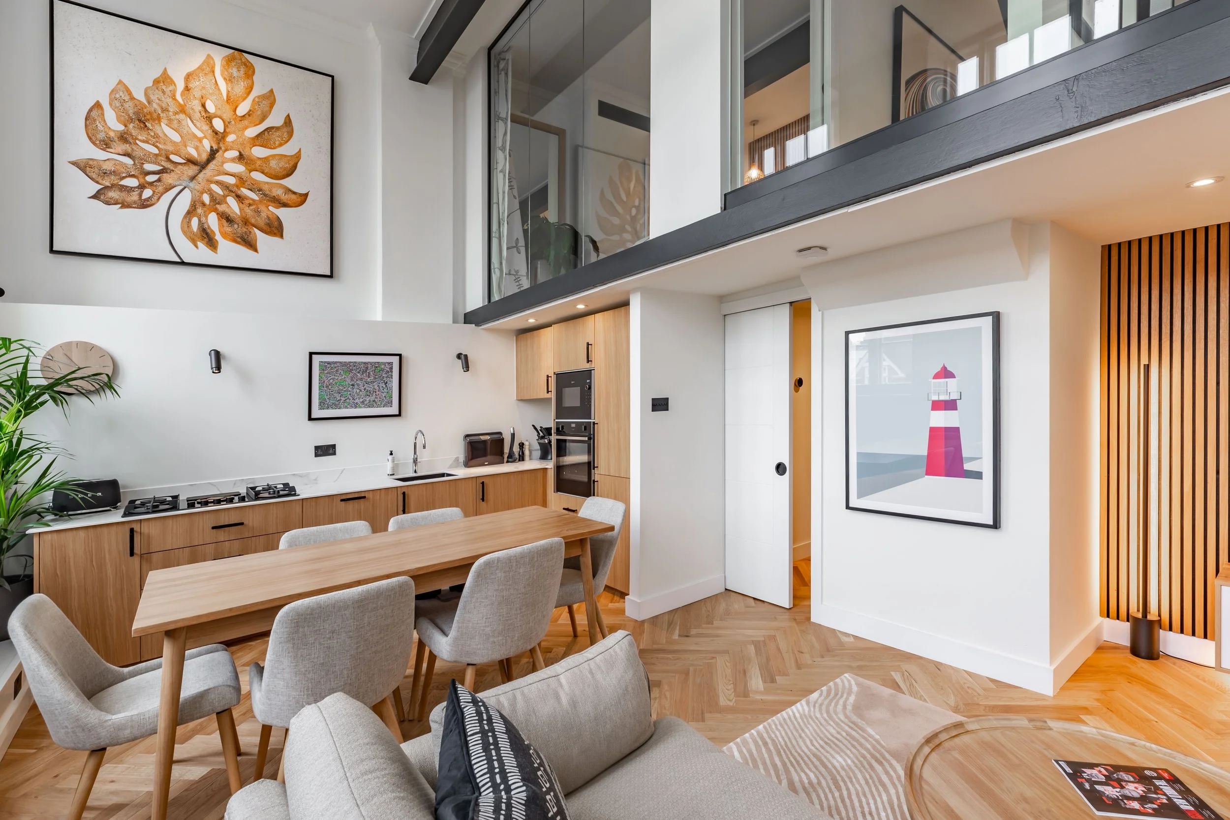 Open-plan living, dining and kitchen in a Grade II listed school conversion London duplex — glass balustrade mezzanine above, pocket door open to herringbone hallway, oak slat feature wall, contemporary furniture. Modern heritage interior design by Studio Akiva