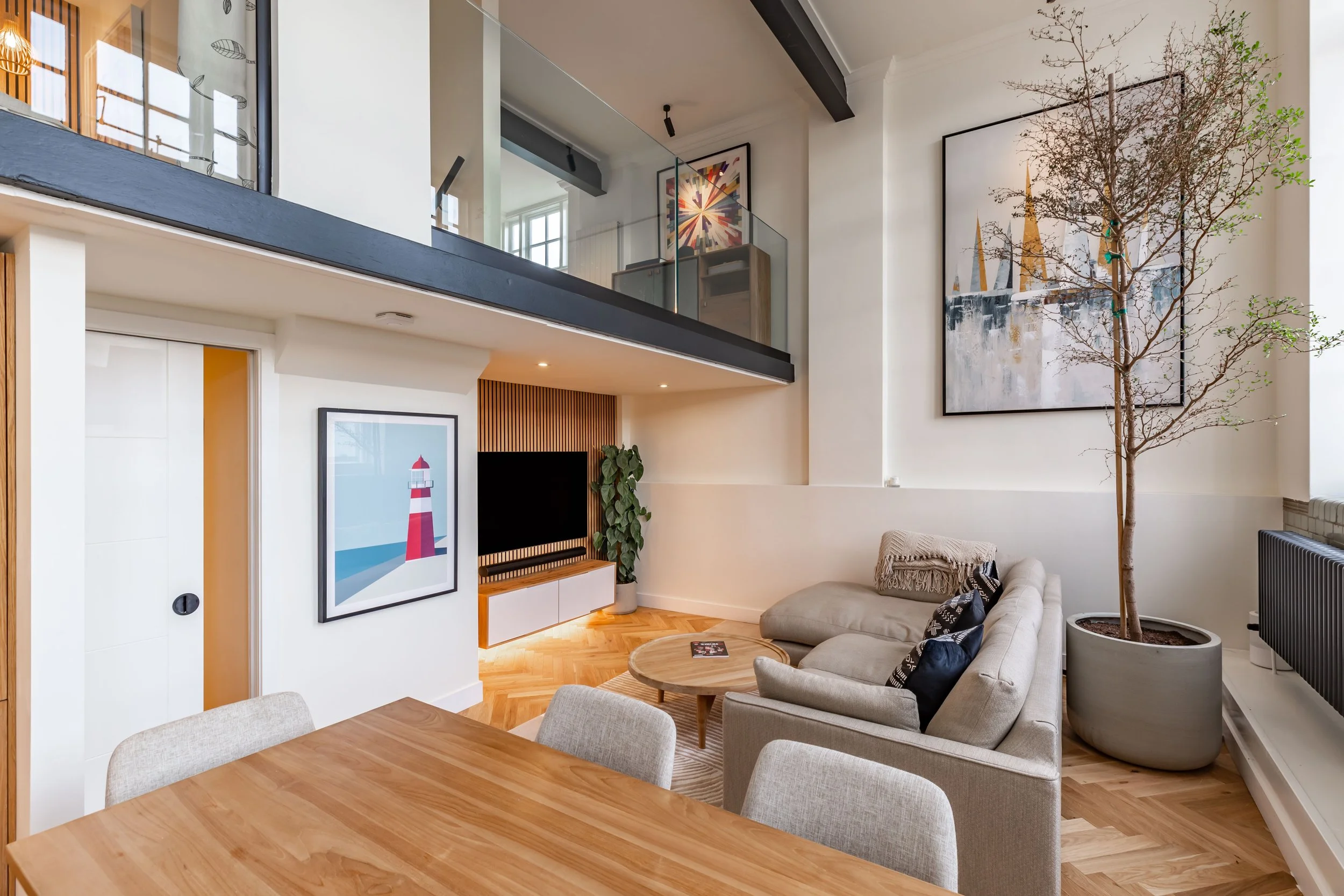 Living room of a London school conversion duplex showing full double-height volume — glass balustrade mezzanine above, oak slat TV wall, grey sofa, sailboat artwork on double-height wall, large indoor tree, herringbone floor. Studio Akiva
