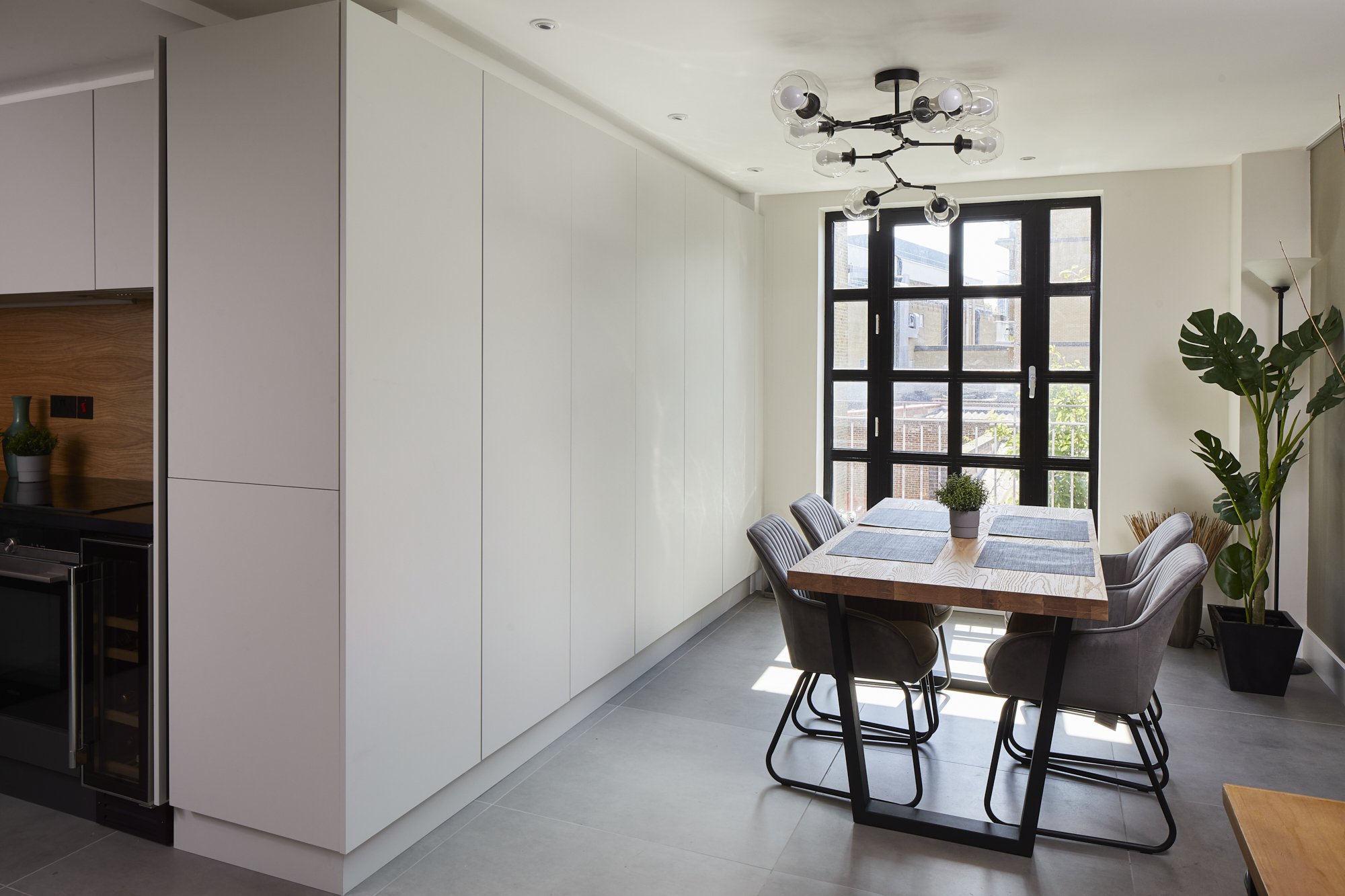 Dining area in a Shoreditch loft renovation — oak dining table, grey upholstered chairs, floor-to-ceiling white storage wall, black-framed grid window with balcony, and black branching chandelier. Design by Studio Akiva