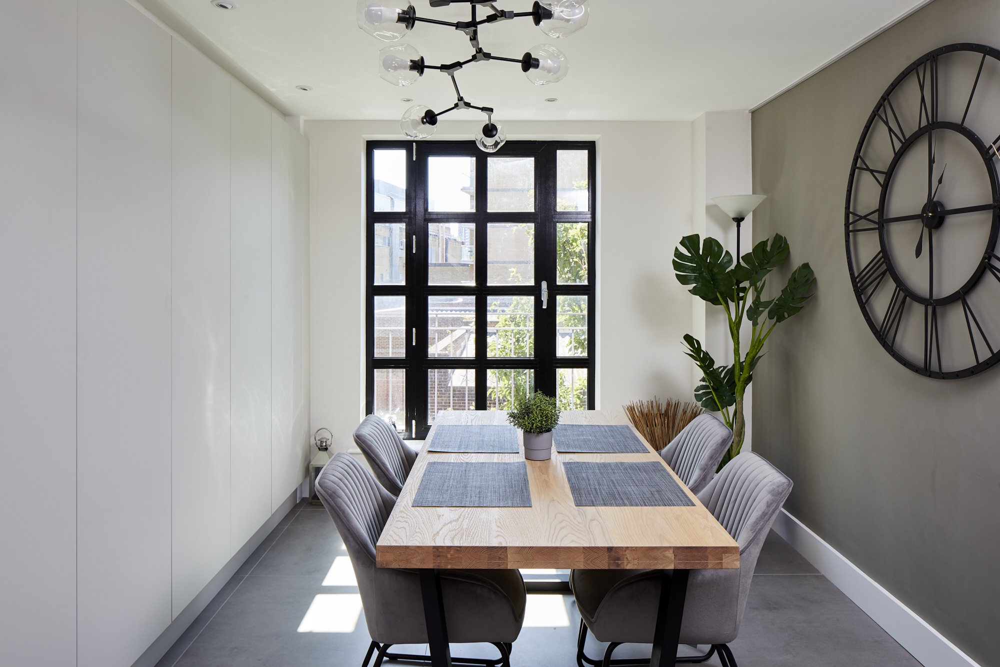 Industrial-style dining room in a Shoreditch loft — full-height black-framed grid window flooded with natural light, oak dining table, grey velvet chairs, oversized black clock, and a khaki accent wall. Studio Akiva, London
