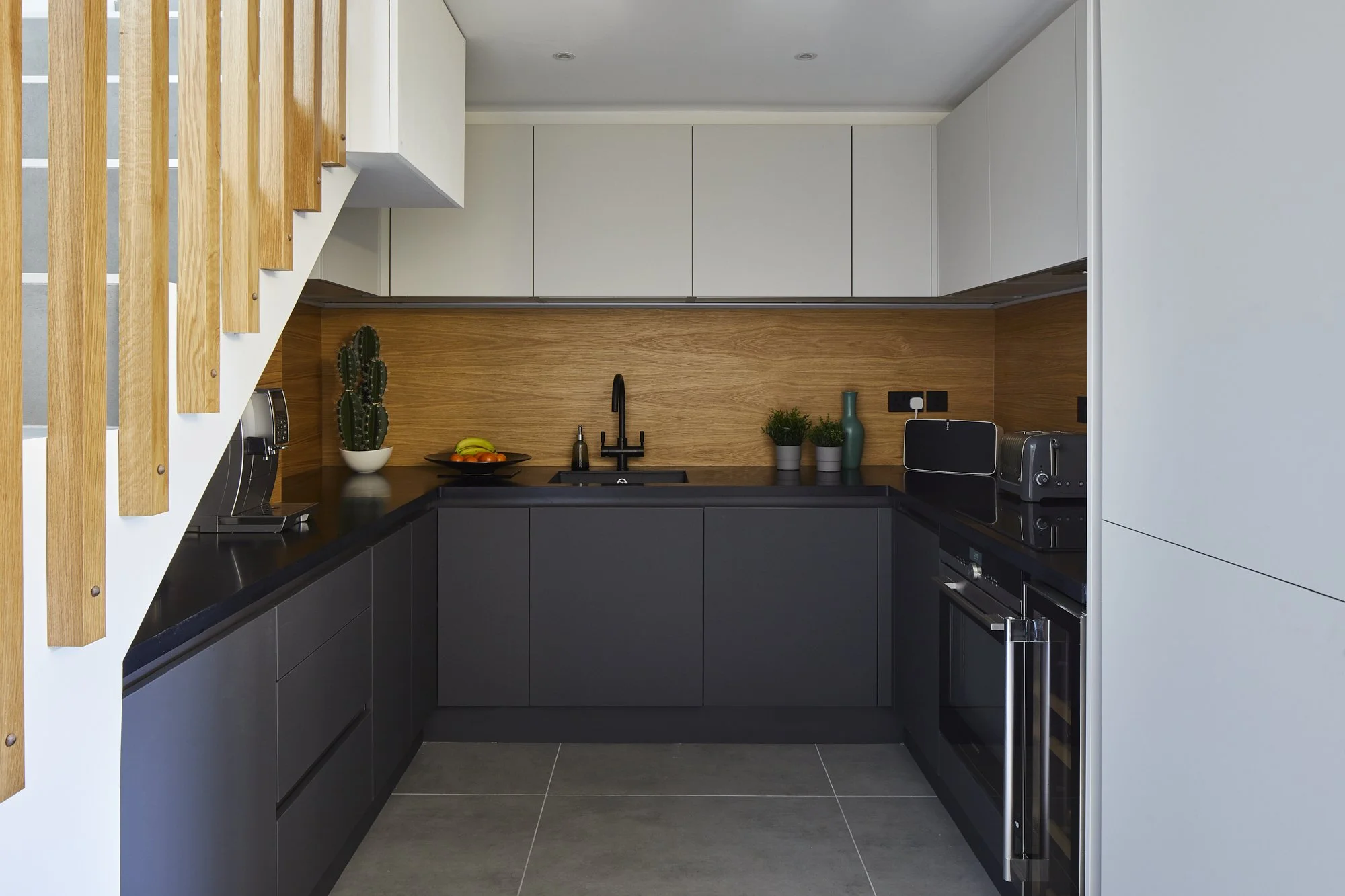 U-shaped industrial kitchen in a Shoreditch loft in daylight — matte black handleless lower cabinets, pale grey upper units, full-height oak splashback and concrete-effect floor tiles. Interior design by Studio Akiva