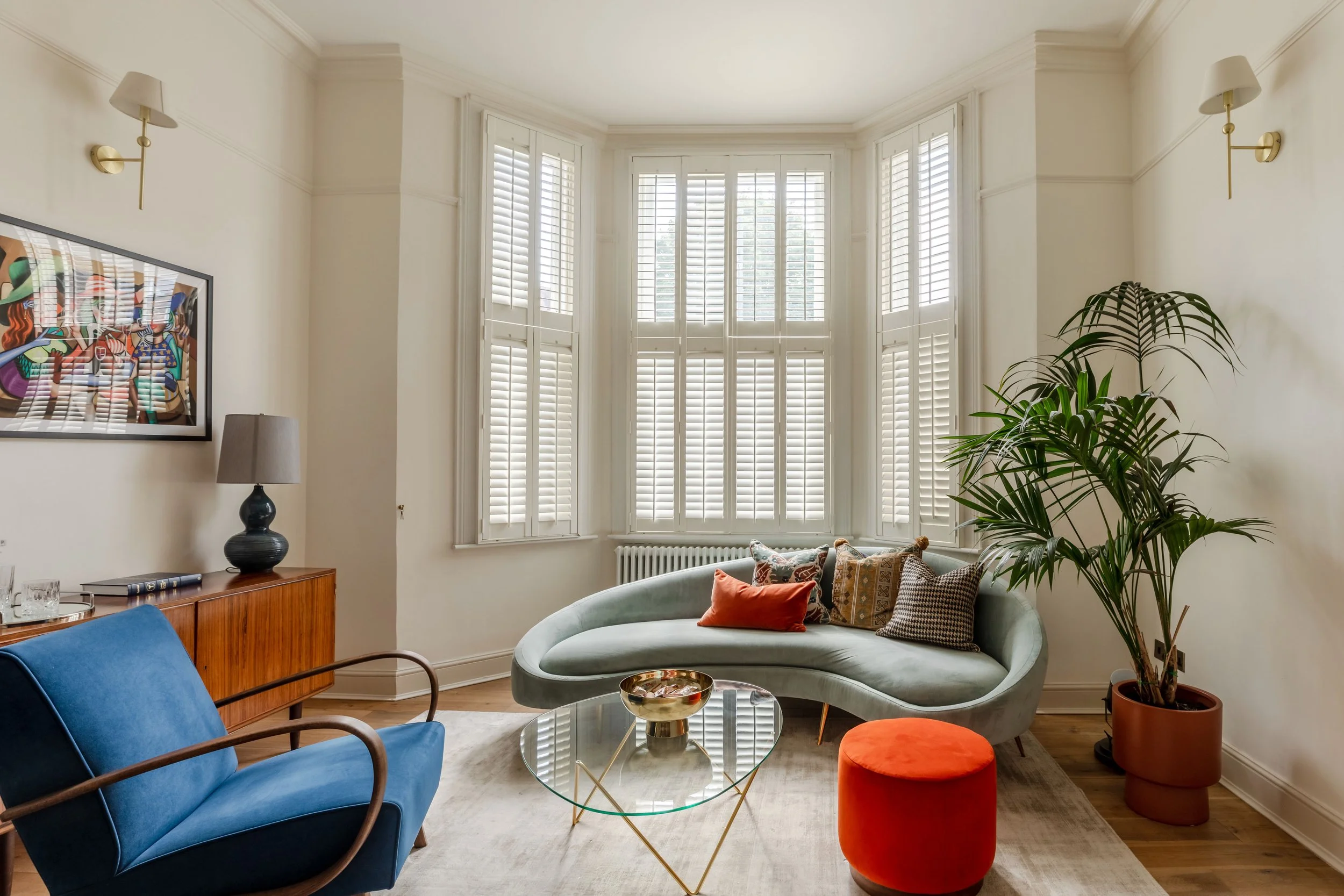 Victorian terraced house living room with plantation shutters on bay windows, curved sage green sofa, blue armchair, midcentury sideboard, and colorful wall art