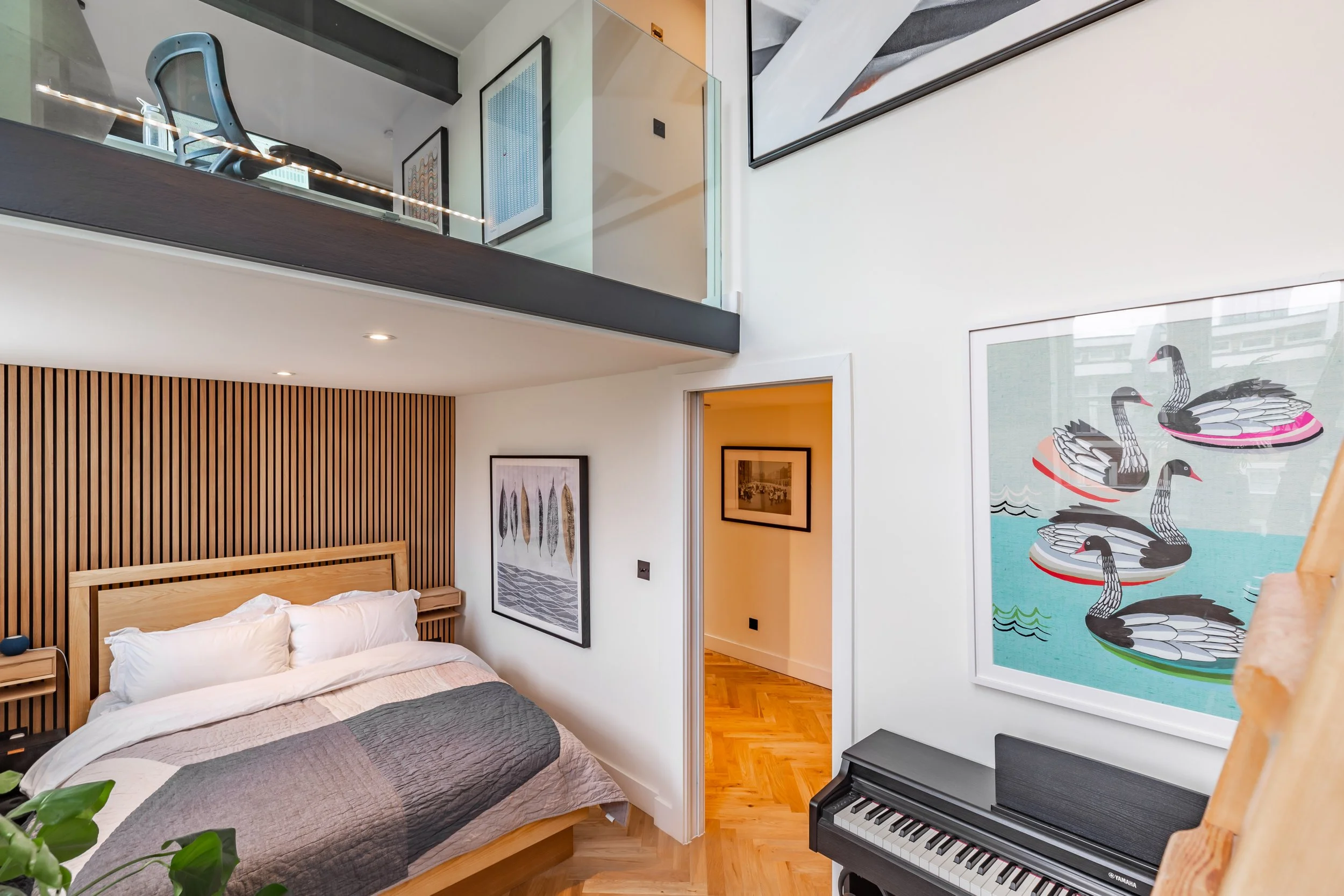 Master bedroom looking back into a converted school duplex London — oak slat wall, oak bed, mezzanine office above with glass balustrade, pocket door open to herringbone hallway, piano in foreground. Studio Akiva interior design
