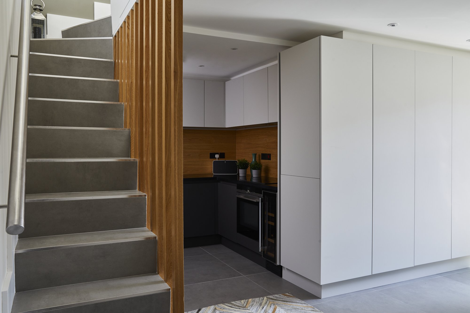 View from the hallway into a Shoreditch industrial loft renovation — concrete-effect stair treads, oak baluster post, full-height pale grey storage wall and kitchen beyond. Interior design by Studio Akiva, London