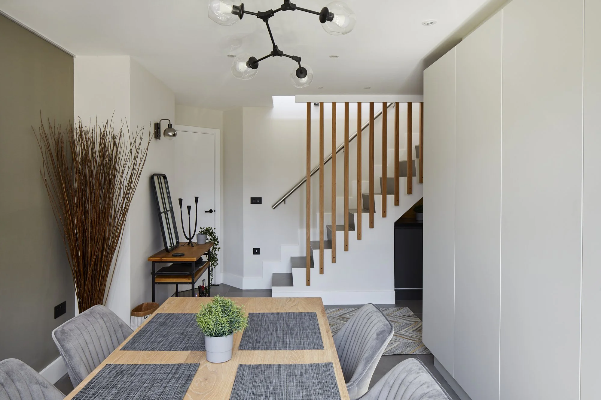 View from the dining table back towards the staircase in a Shoreditch loft — branching black chandelier, grey accent wall, oak baluster staircase and pale full-height storage. Interior design by Studio Akiva