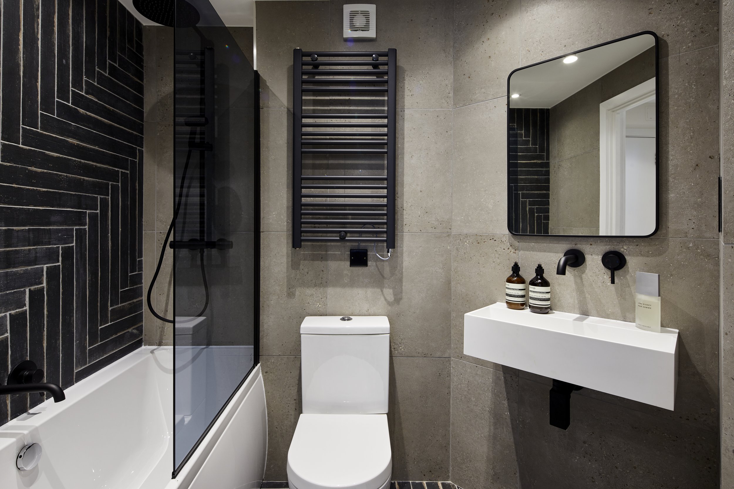 After photo — industrial main bathroom in a Shoreditch loft renovation with large-format concrete-effect grey tiles, dark herringbone black tiles on the bath wall, matte black heated towel rail, wall-hung white basin, black-framed mirror and matte black fittings throughout. Design by Studio Akiva, London