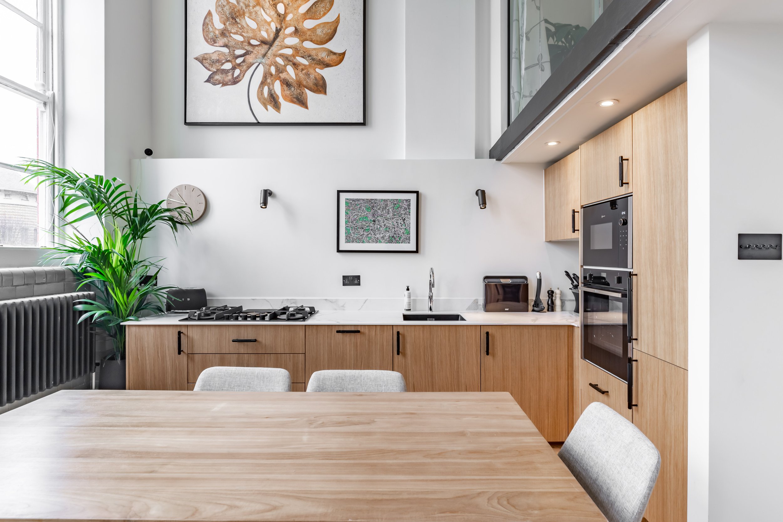 Kitchen in a Grade II listed school conversion duplex London — oak-effect cabinets with matte black bar handles, white Calacatta quartz worktop and splashback, matte black gas hob, Neff oven tower, wall sconces, original column radiator. Studio Akiva interior design