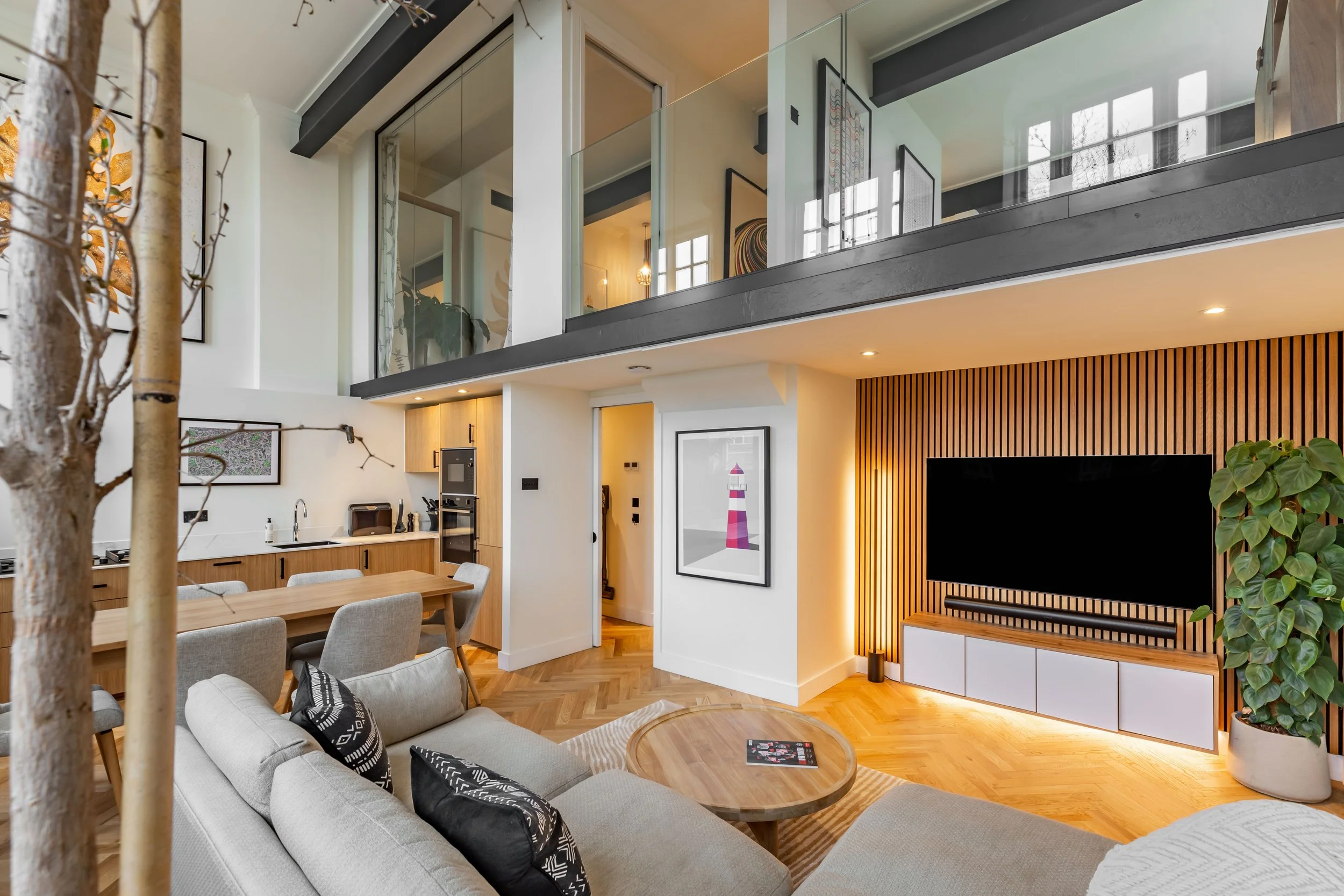Evening view of a Grade II listed school conversion duplex in London — oak slat TV wall with warm amber backlighting, mezzanine bedroom warmly lit above with pendant lights visible through glass balustrade, kitchen and dining beyond. Interior design by Studio Akiva