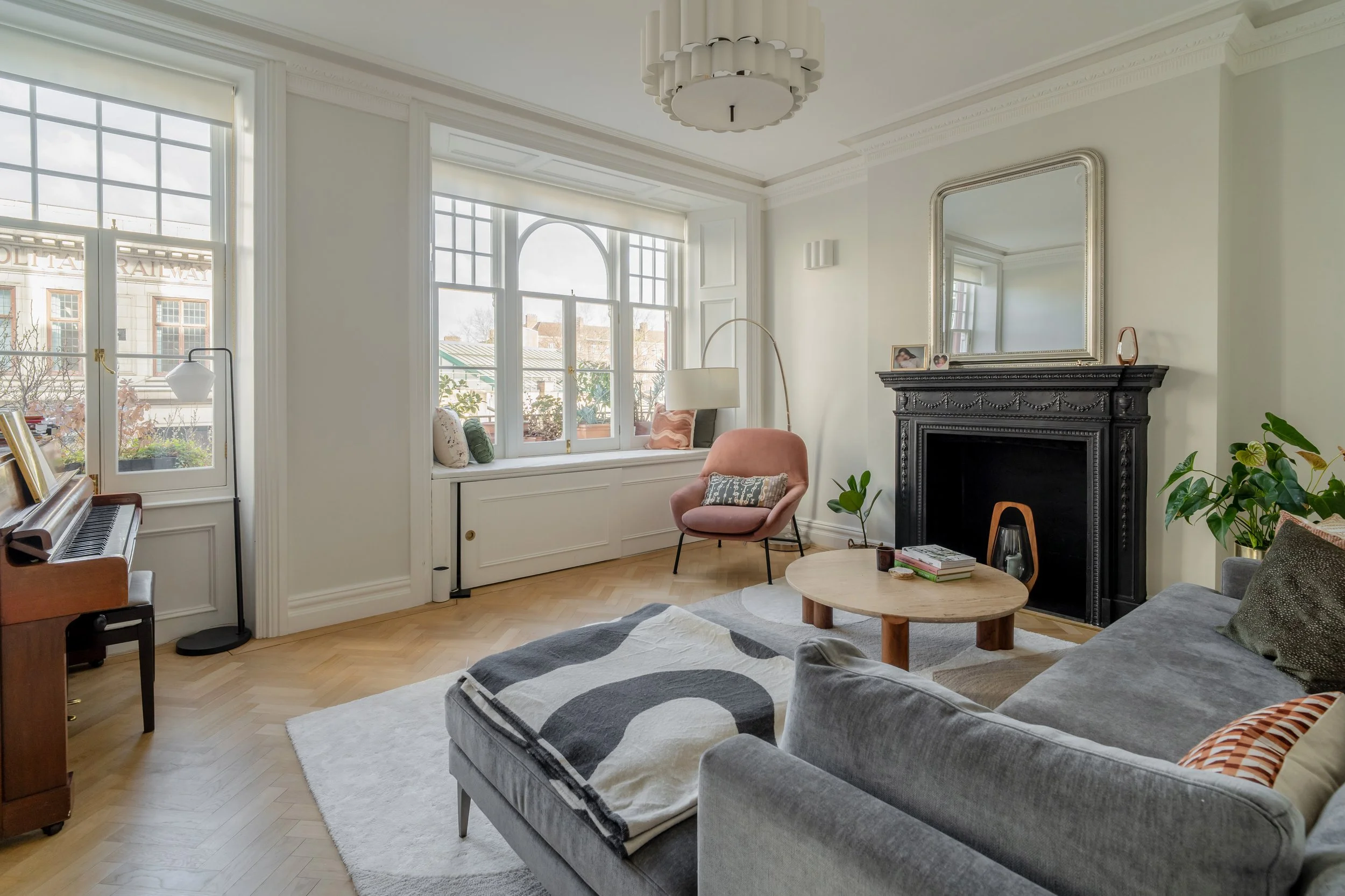 Bright contemporary living room with herringbone flooring, statement black fireplace, large sash windows, and soft neutral furnishings.