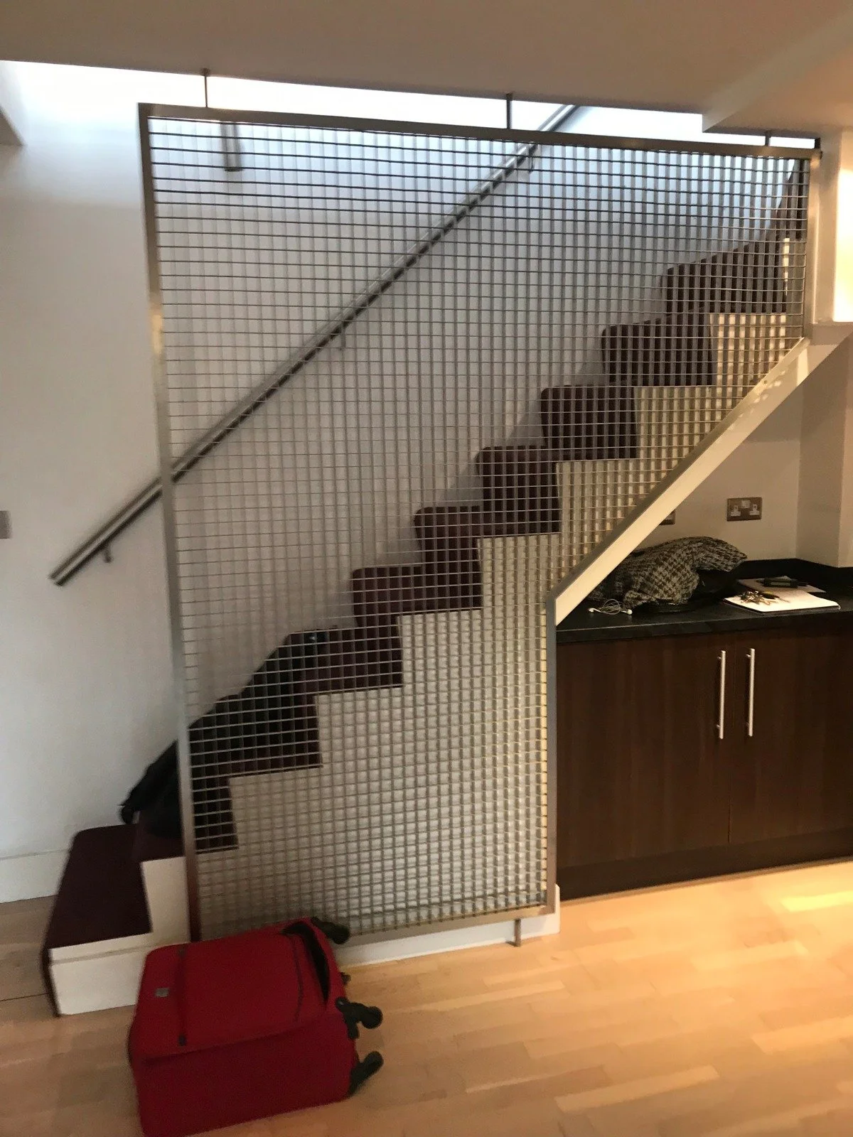 Before renovation photo of a Shoreditch loft staircase with outdated metal grid balustrade, laminate flooring, and a red suitcase on the floor