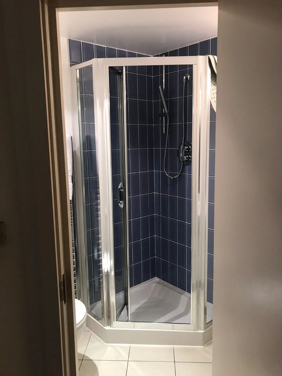 Before renovation photo of an en-suite bathroom with a dated corner shower cubicle, blue tile walls, chrome fittings, and white floor tiles