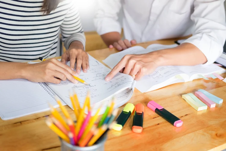 Two people reviewing notes together at a desk with pencils and highlighters.