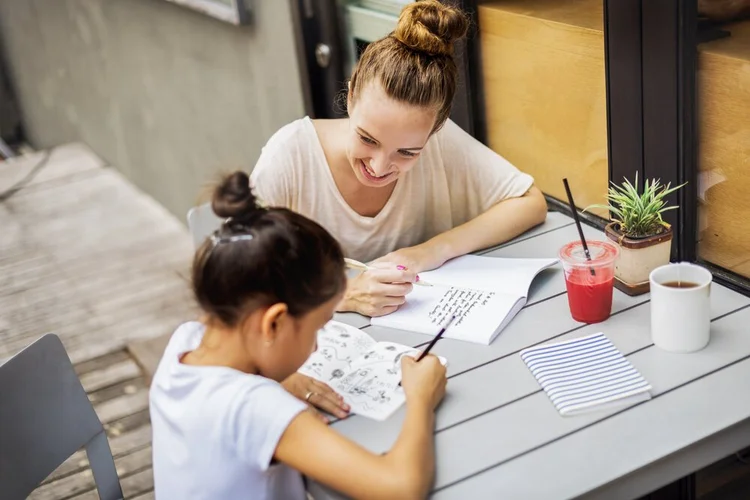 Woman helping a child with drawing at an outdoor table.