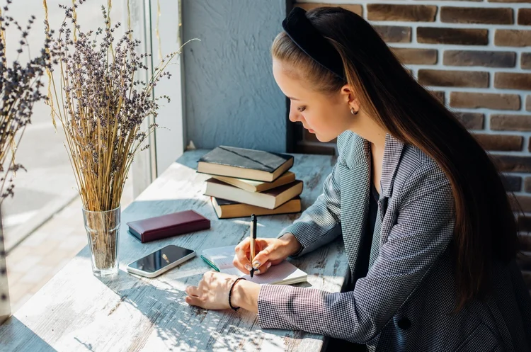 Woman writing in a notebook at a desk by the window, with books and a phone nearby.