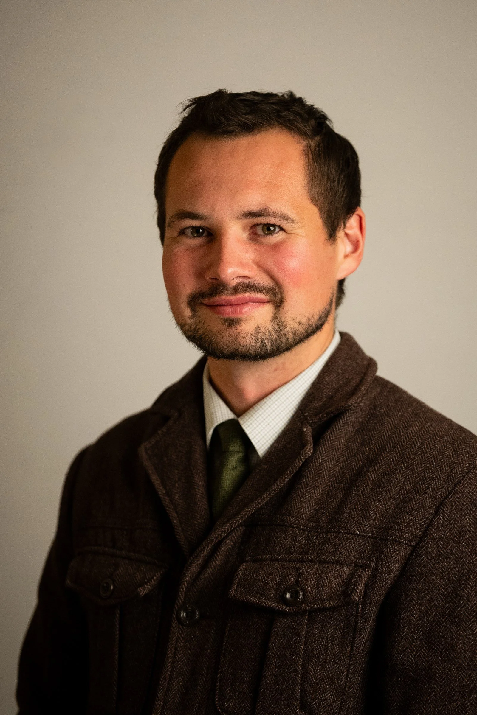 Studio headshot of a man in a brown jacket and tie against a light background in Potsdam, NY