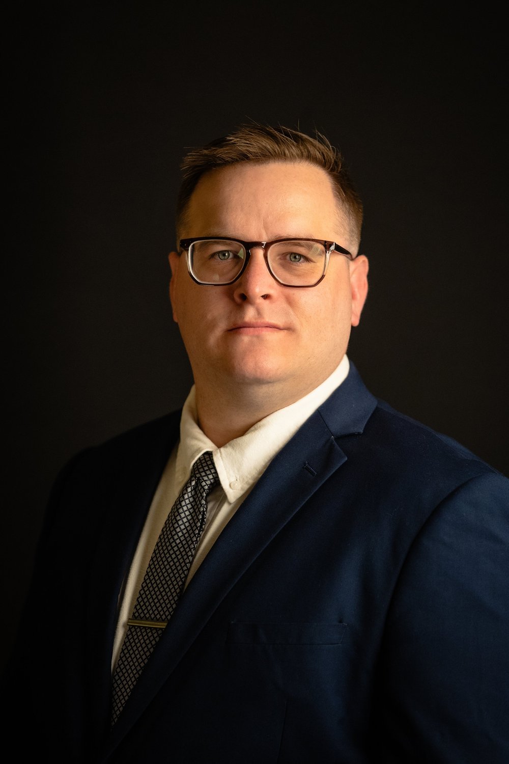 Studio headshot of a man in glasses, a navy suit and tie against a dark background, photographed by JD Meyers Productions in Potsdam