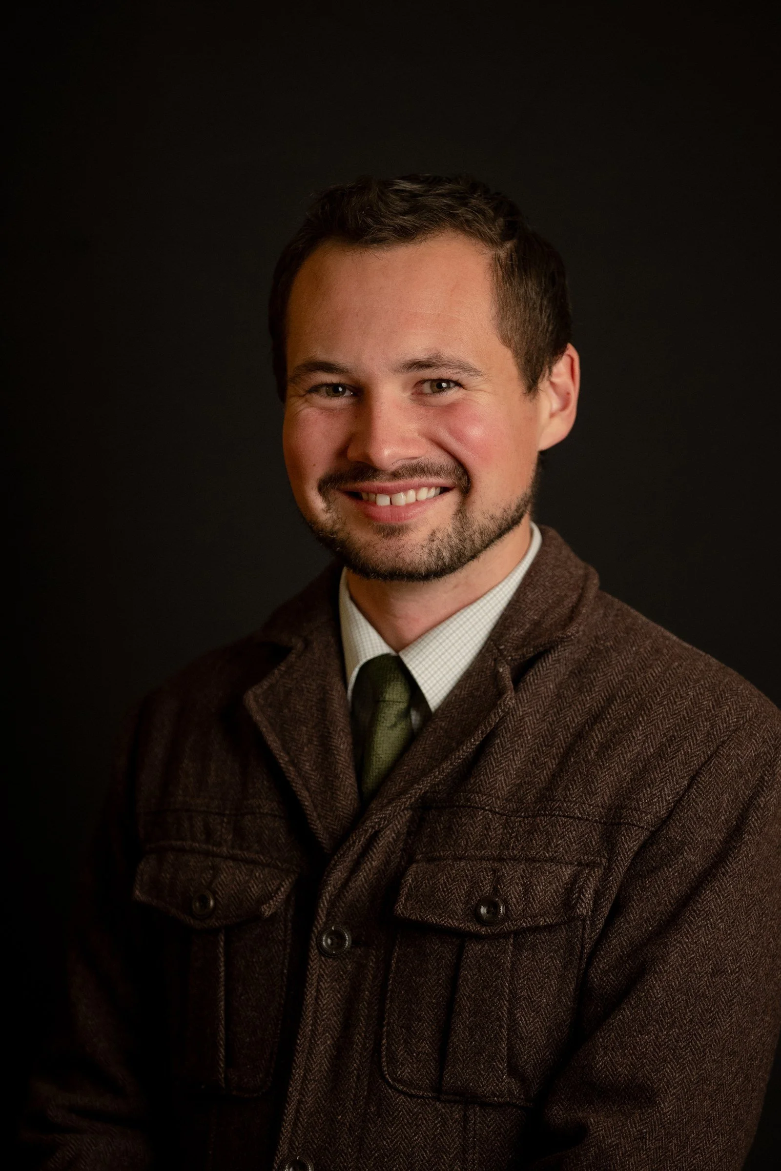 Smiling studio headshot of a man in a brown jacket and tie against a dark background in Potsdam, NY