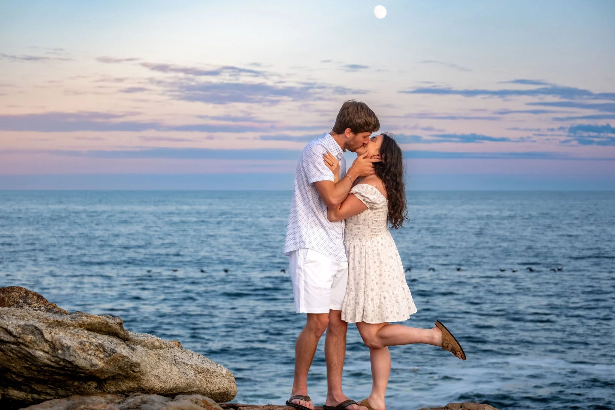 Kirstie kicking one foot up as Lucas kisses her on the rocks with the moon over the ocean at Reid State Park in Georgetown Maine engagement photography VMG Productions