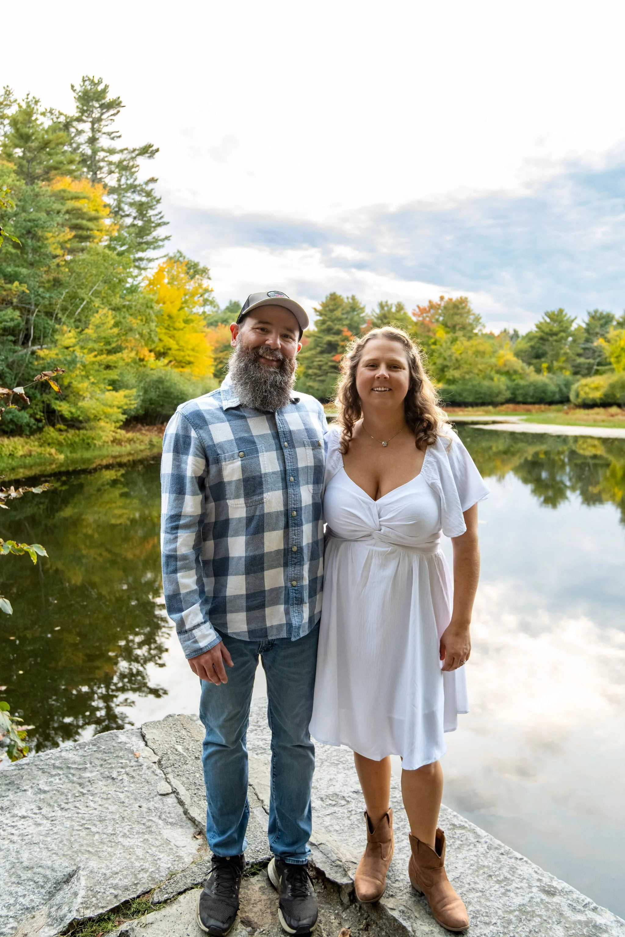 Alison and Dave standing on the granite blocks at the edge of the pond with the colored shoreline behind them at Vaughan Woods in Hallowell Maine fall engagement photography VMG Productions