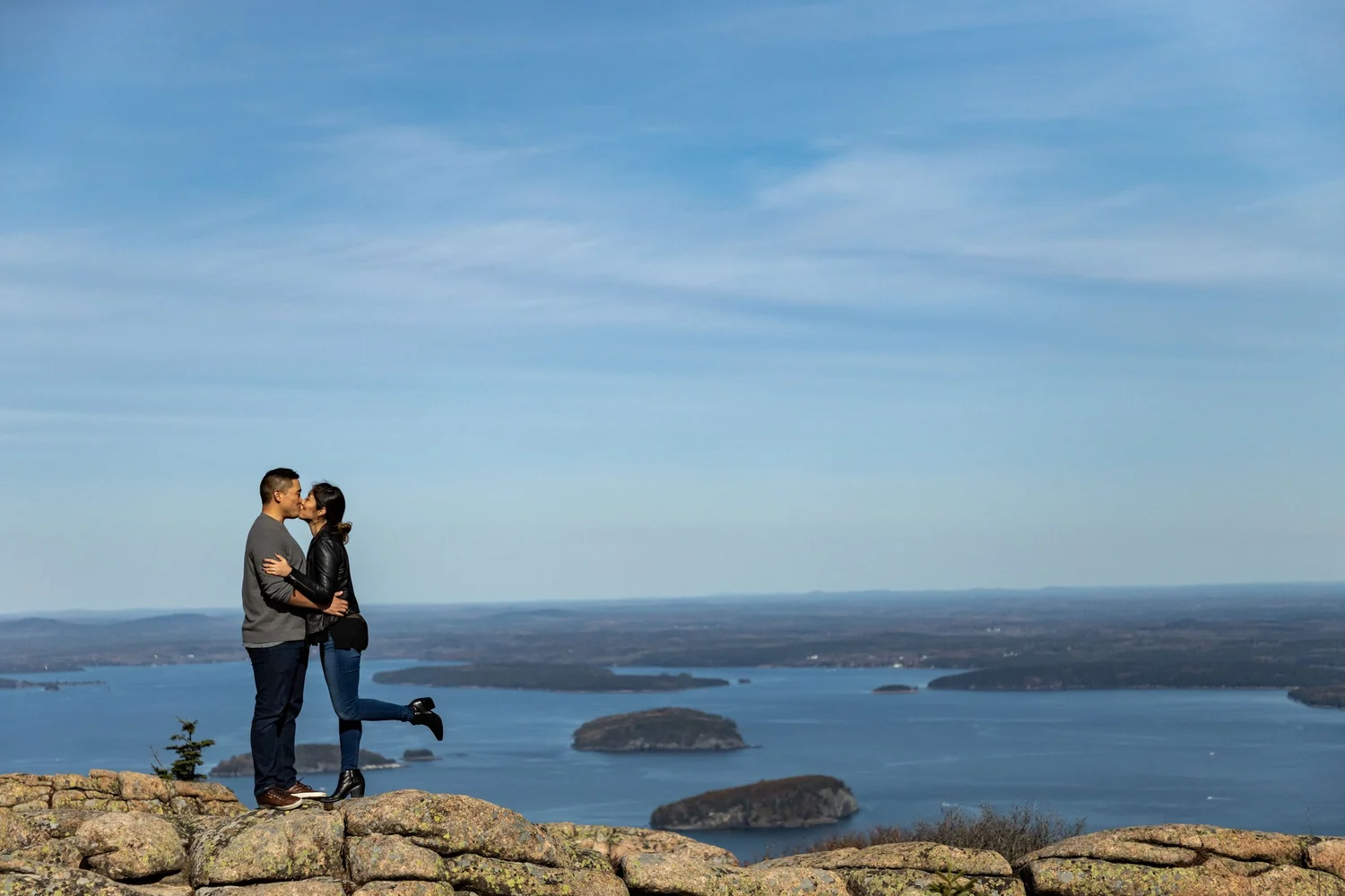 Agnes and Peter kissing on Cadillac Mountain summit wide shot ocean and islands behind Acadia National Park engagement VMG Productions