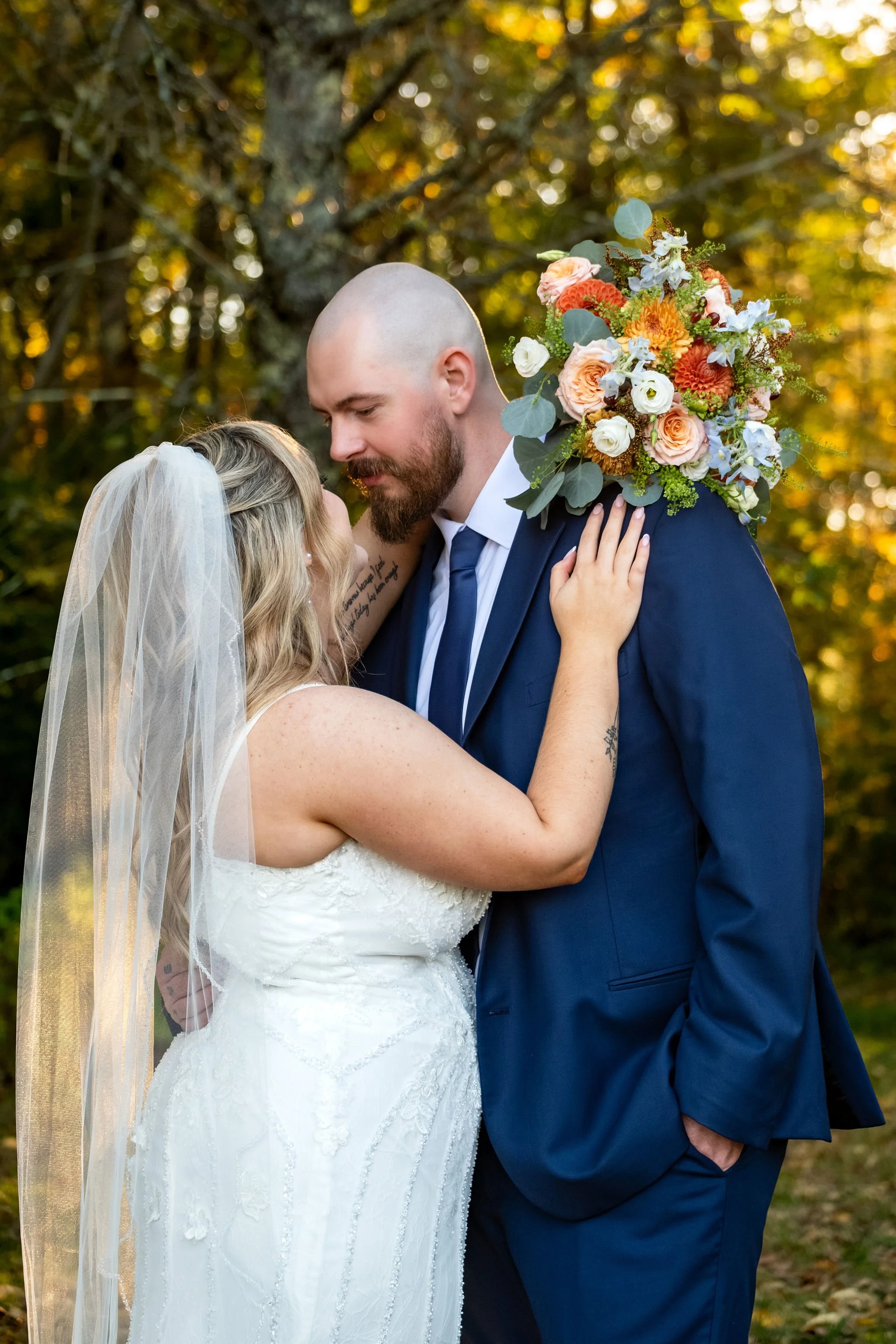 Ellie resting her hand on Sam's chest as he kisses her head in the golden woods at Boothbay Playhouse in Boothbay Maine fall wedding photography VMG Productions