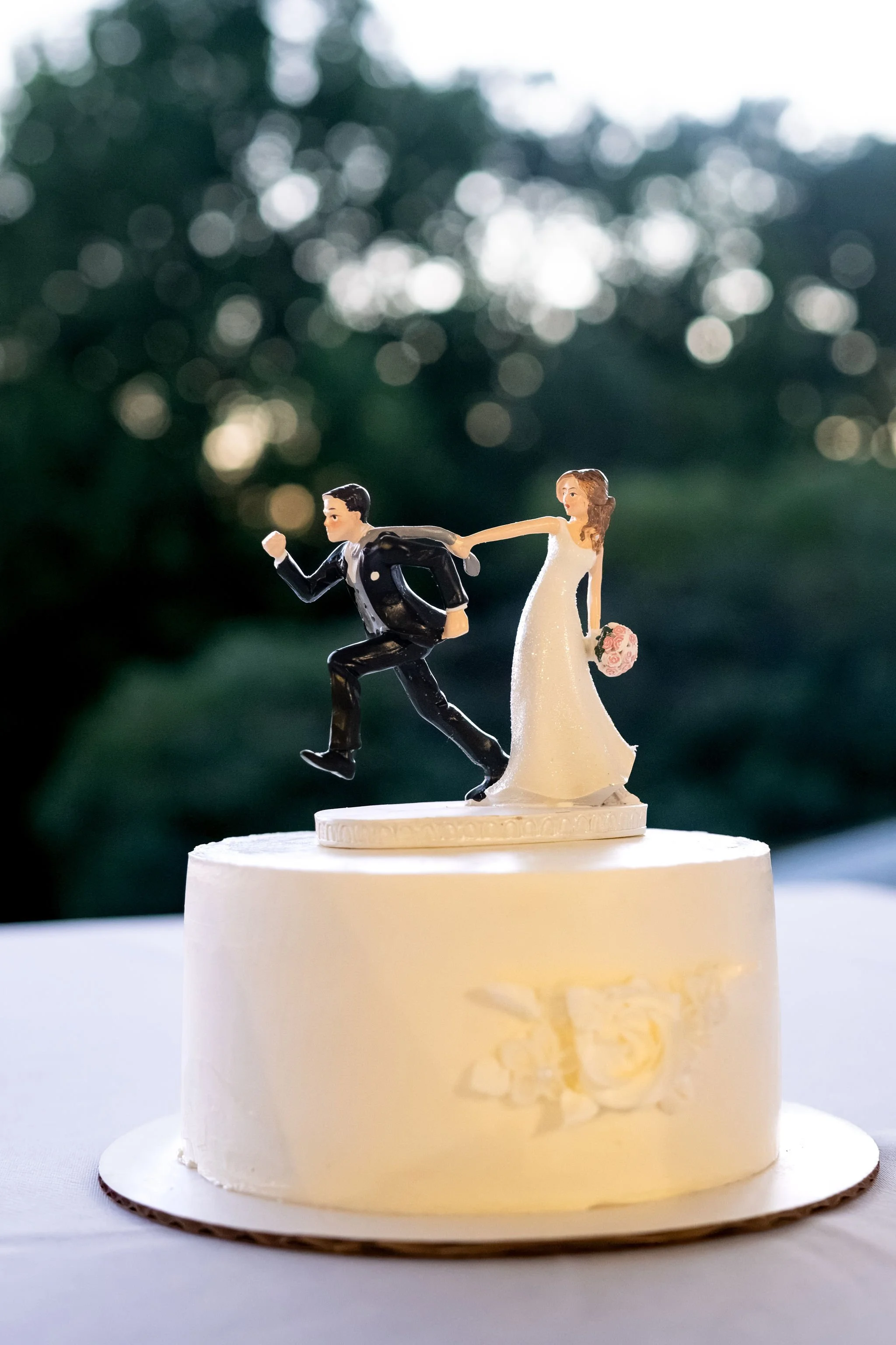 White wedding cake with a topper of the bride dragging a running groom back, with the trees soft behind it at The Lucerne Inn in Dedham Maine summer wedding photography VMG Productions