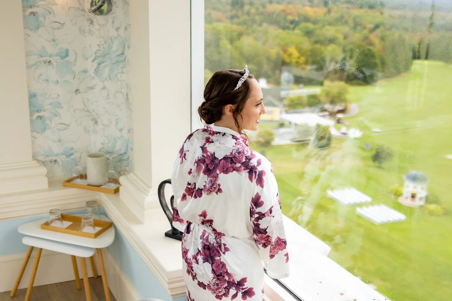 Bride in floral robe looking out window at ceremony setup below at Mountain View Grand Resort Whitefield New Hampshire wedding morning
