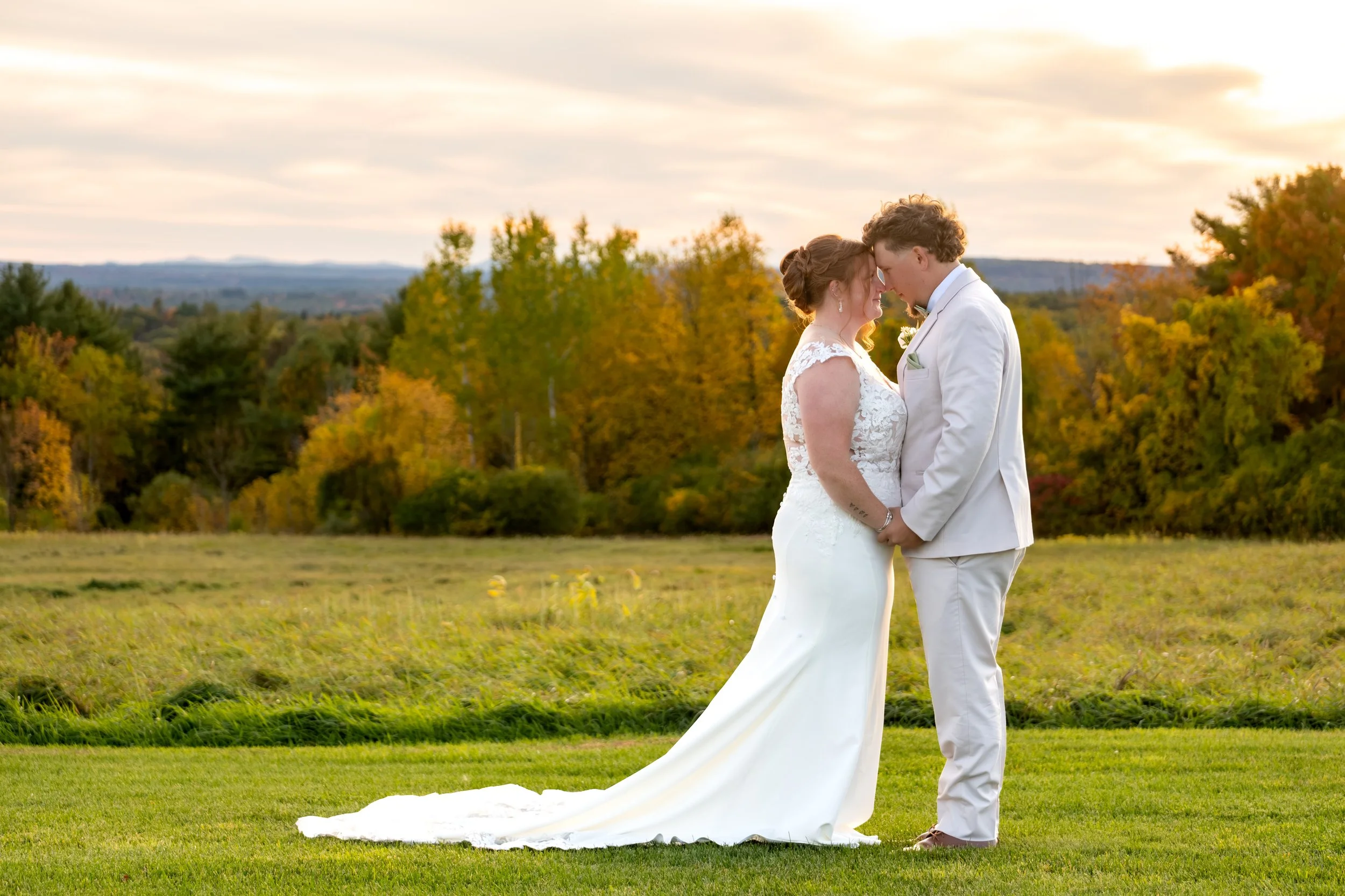 Andrew and Erika kissing in the green field with fall trees behind them at the Maine Wedding Barn and Event Center in Minot Maine fall wedding photography VMG Productions
