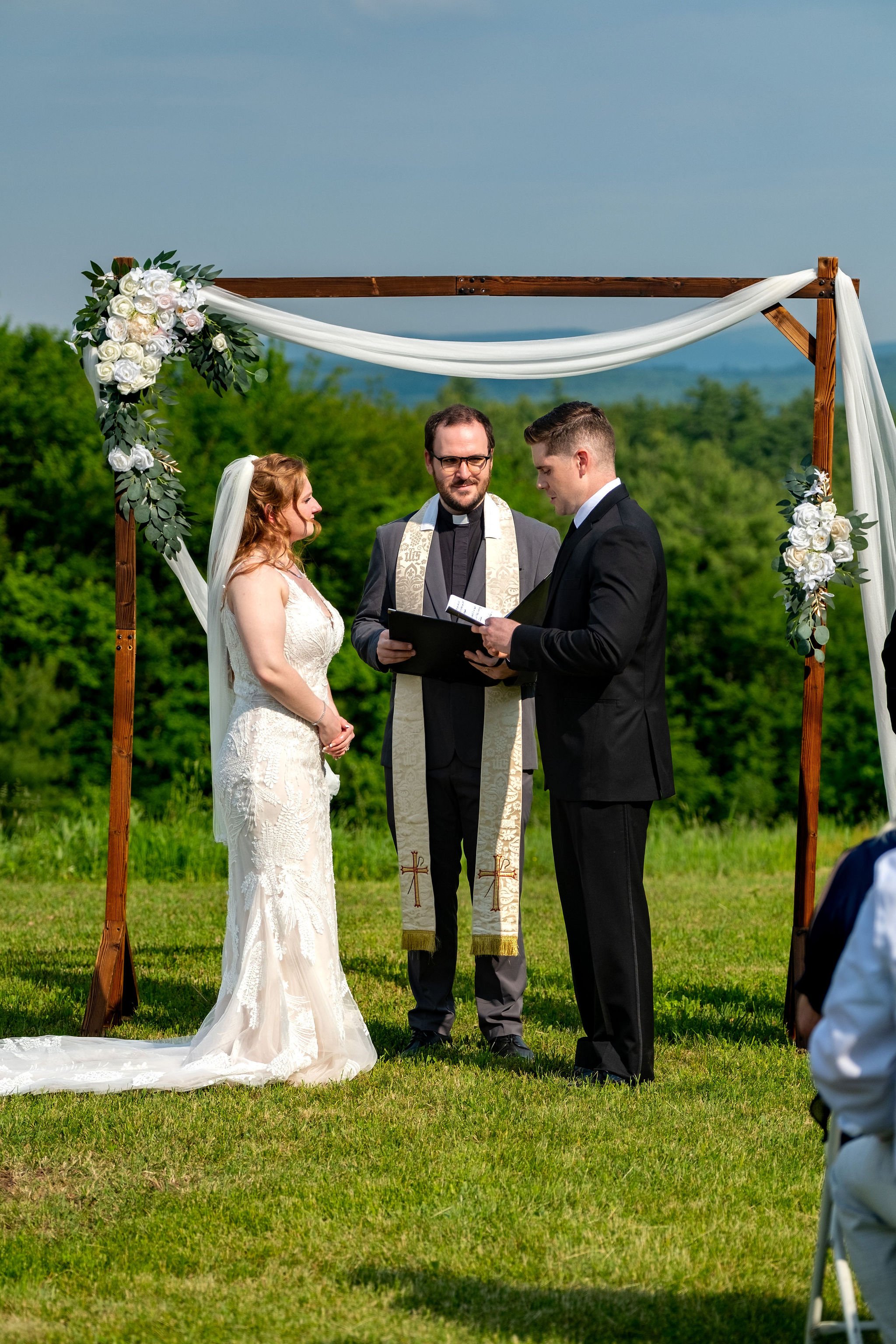 Kayla and Ethan DeAngelis exchanging vows under the wooden arch with the New Hampshire hills behind them at their Sanbornton backyard wedding photography VMG Productions