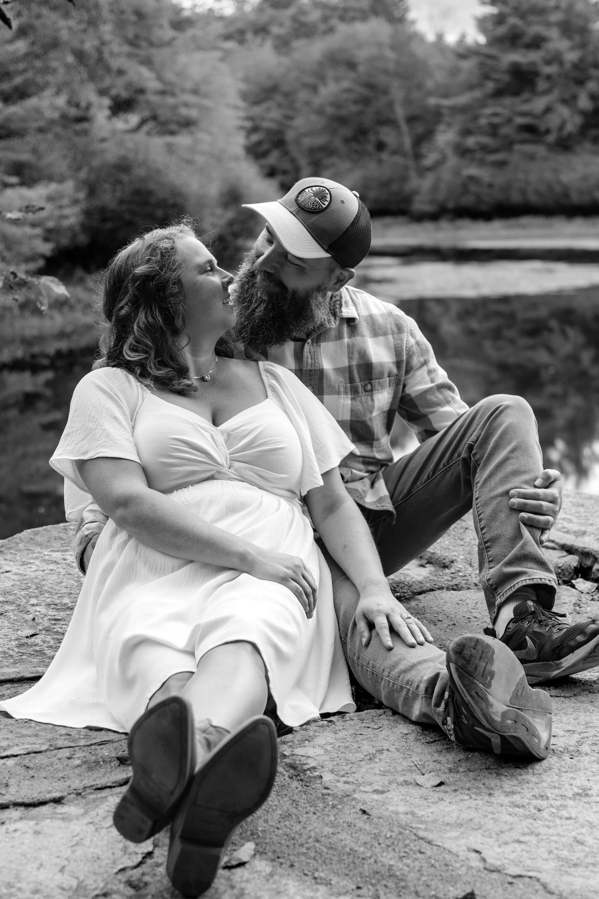 Black and white of Alison and Dave sitting close on the granite at the pond at Vaughan Woods in Hallowell Maine fall engagement photography VMG Productions