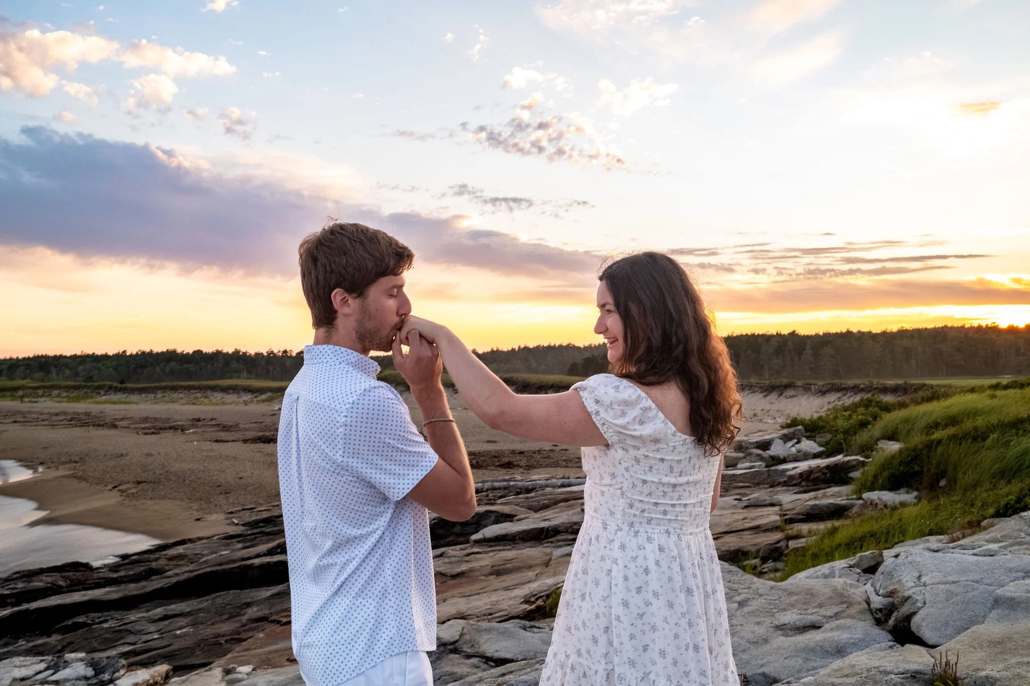 Lucas kissing Kirstie's hand on the rocks with the sunset sky behind them at Reid State Park in Georgetown Maine engagement photography VMG Productions