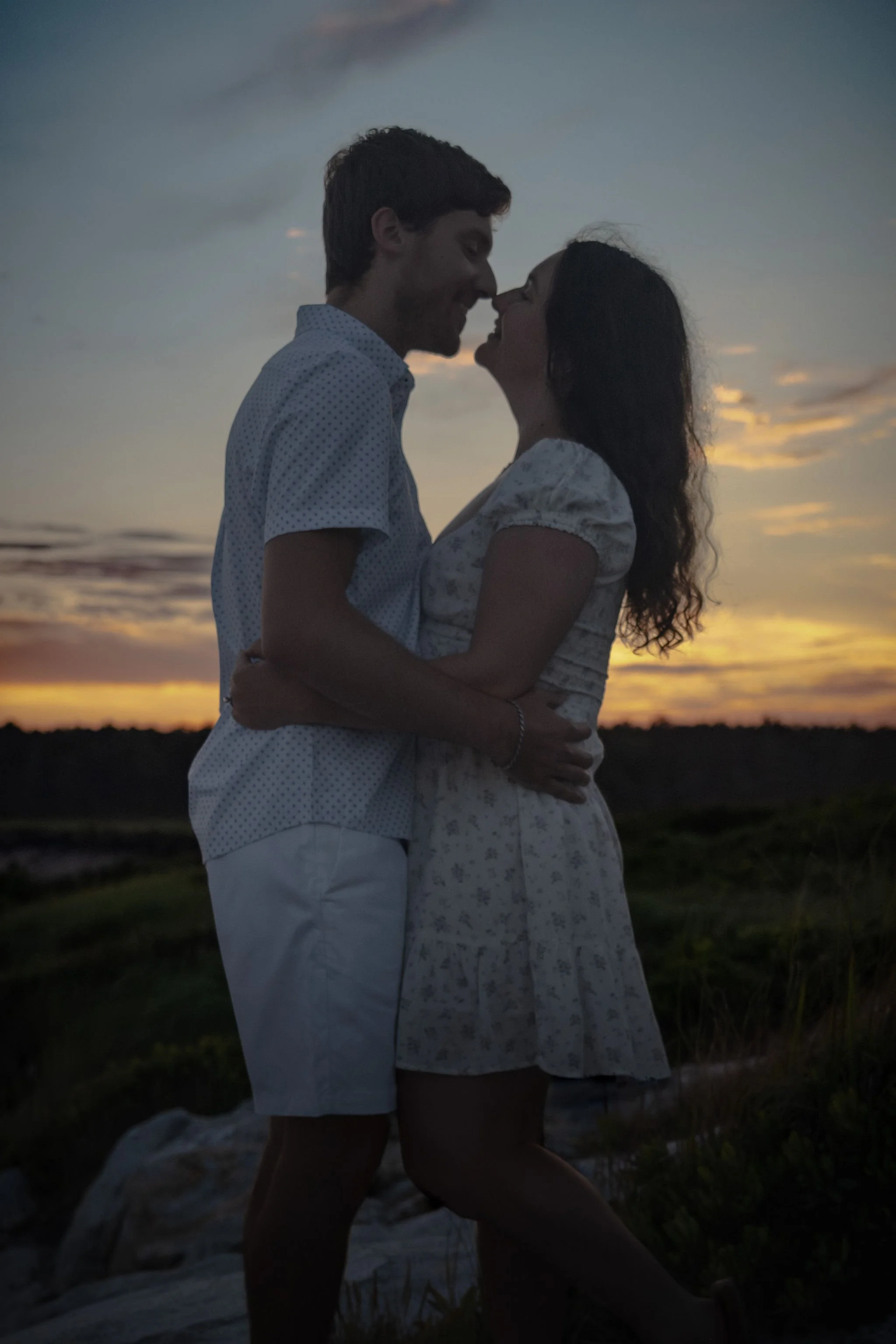 Kirstie and Lucas kissing on the rocks with the last violet light over the marsh at Reid State Park in Georgetown Maine engagement photography VMG Productions
