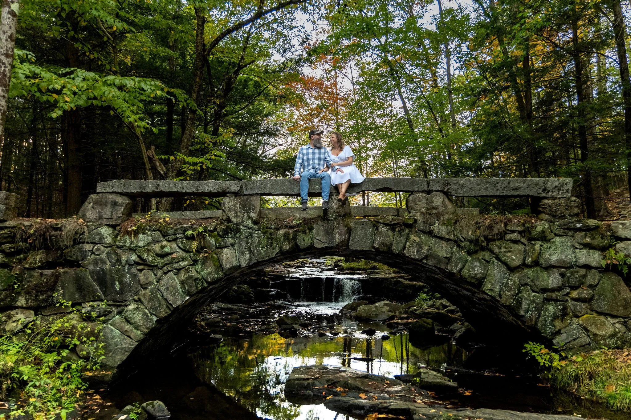 Wide view of the stone arch bridge with Alison and Dave sitting together on the wall and the brook reflecting the trees below at Vaughan Woods in Hallowell Maine fall engagement photography VMG Productions