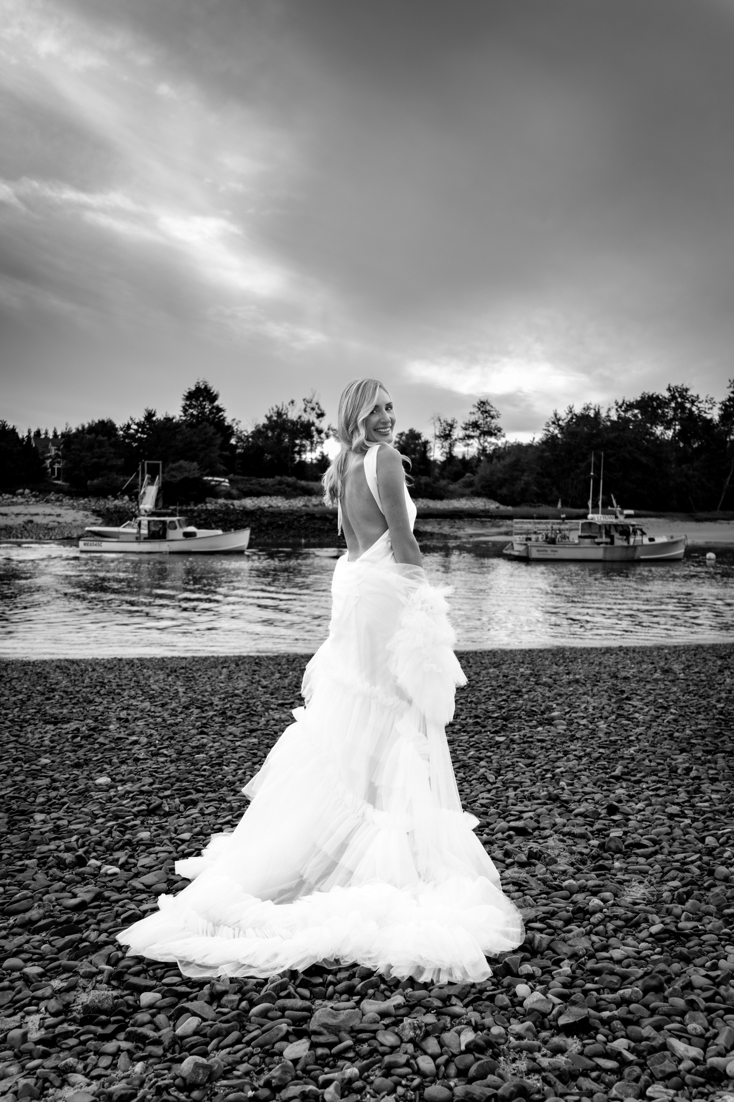 Ashley Draper looking back over her shoulder beneath a dramatic evening sky with lobster boats behind her in black and white at The Breakwater Inn in Kennebunkport Maine summer wedding photography VMG Productions