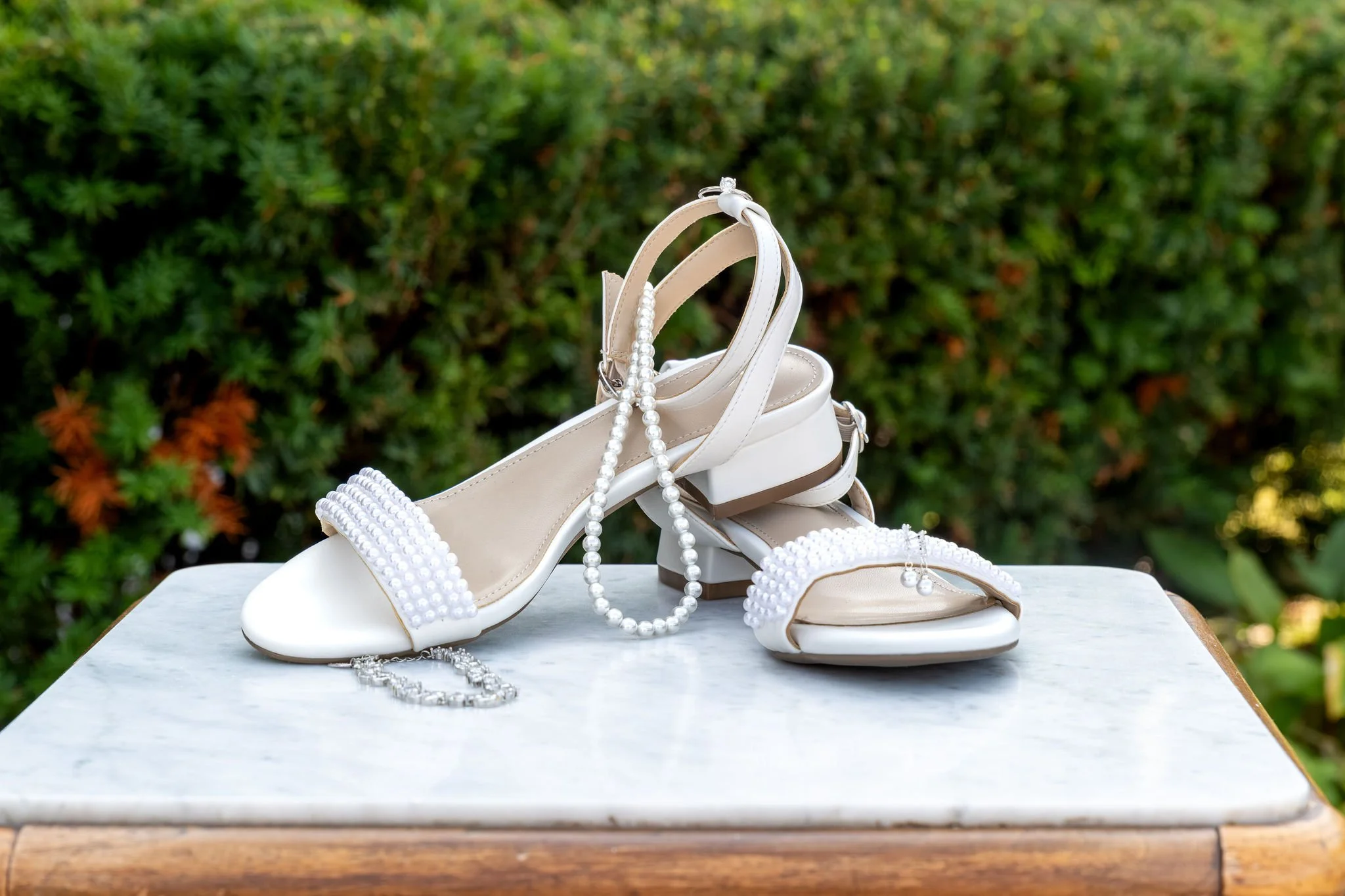 Hollie's white pearl-strap block heel sandals with a pearl necklace and bracelet on a marble table in the garden at The Lucerne Inn in Dedham Maine summer wedding photography VMG Productions