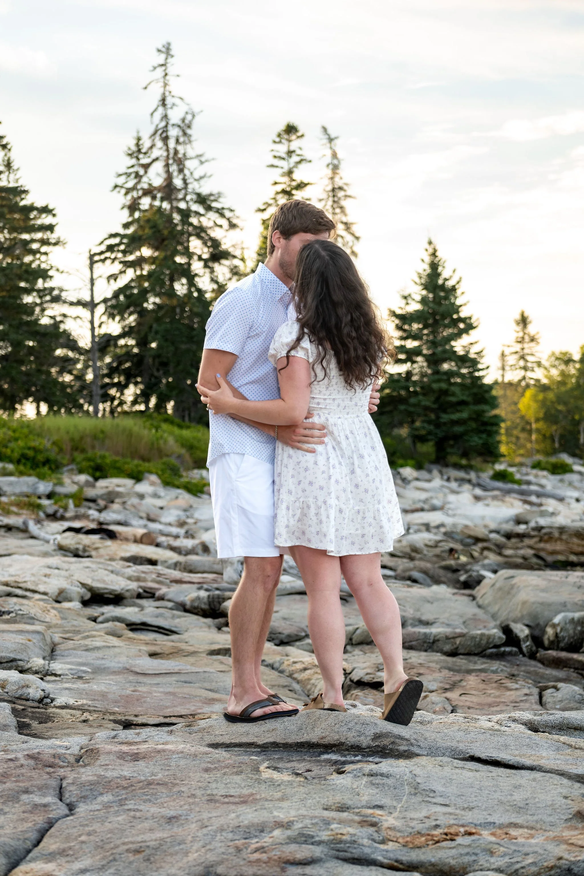 Lucas kissing Kirstie from behind as they stand together on the ledges under the spruce at Reid State Park in Georgetown Maine engagement photography VMG Productions