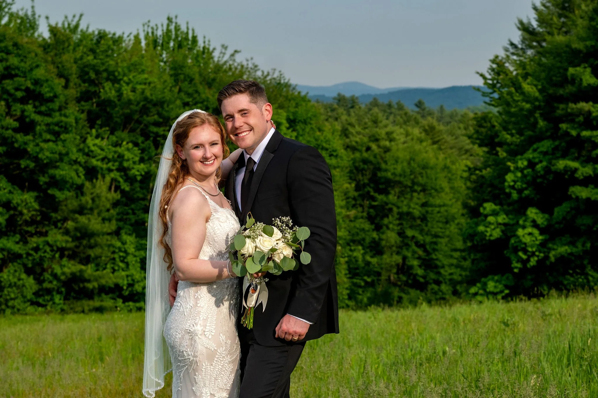 Kayla and Ethan DeAngelis walking arm in arm through the rolling field with the New Hampshire hills behind them at their Sanbornton wedding photography VMG Productions