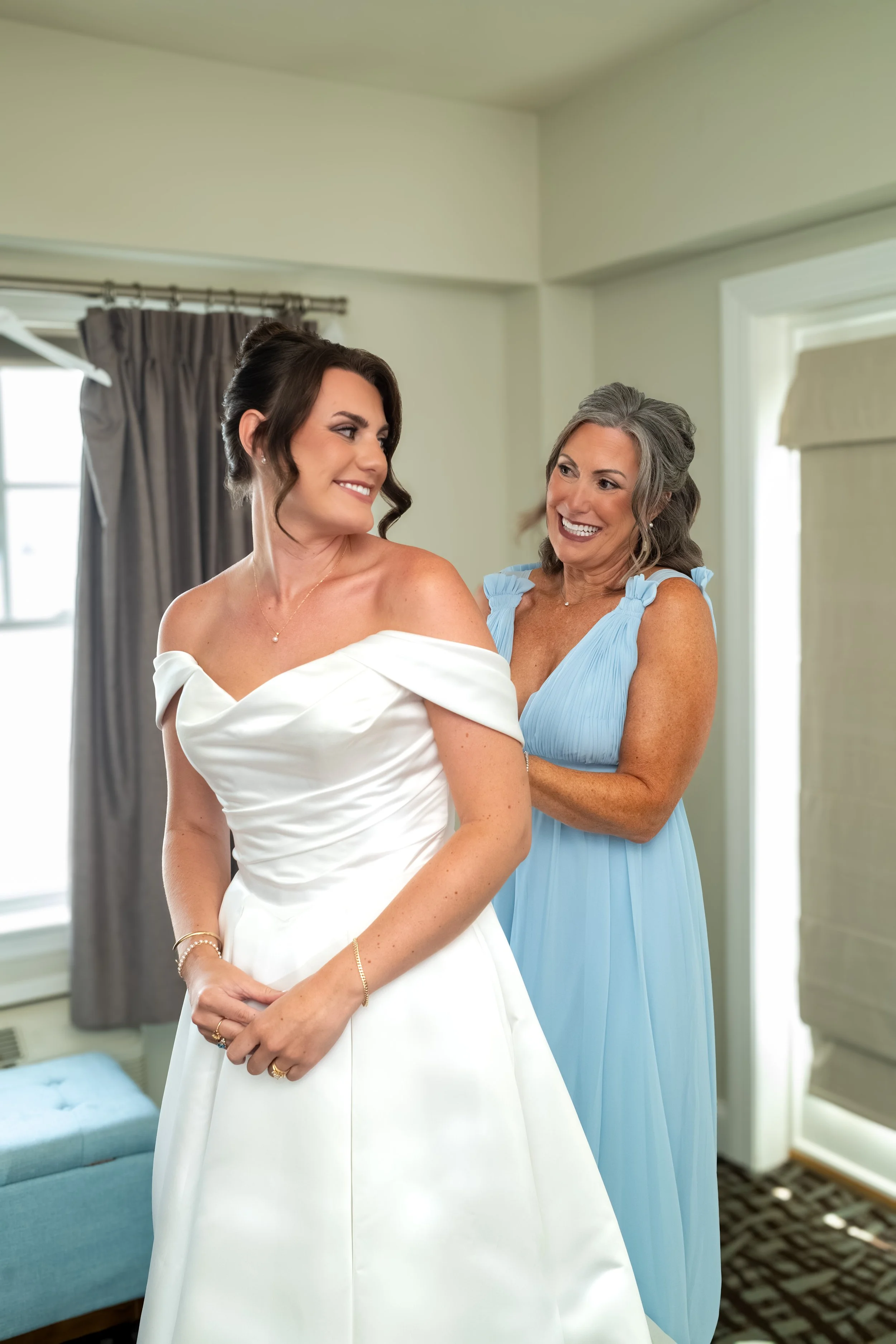 Caroline Pirolli in her gown smiling at her mother Shelly in light blue as she buttons the dress at Union Bluff Hotel in York Maine wedding photography VMG Productions