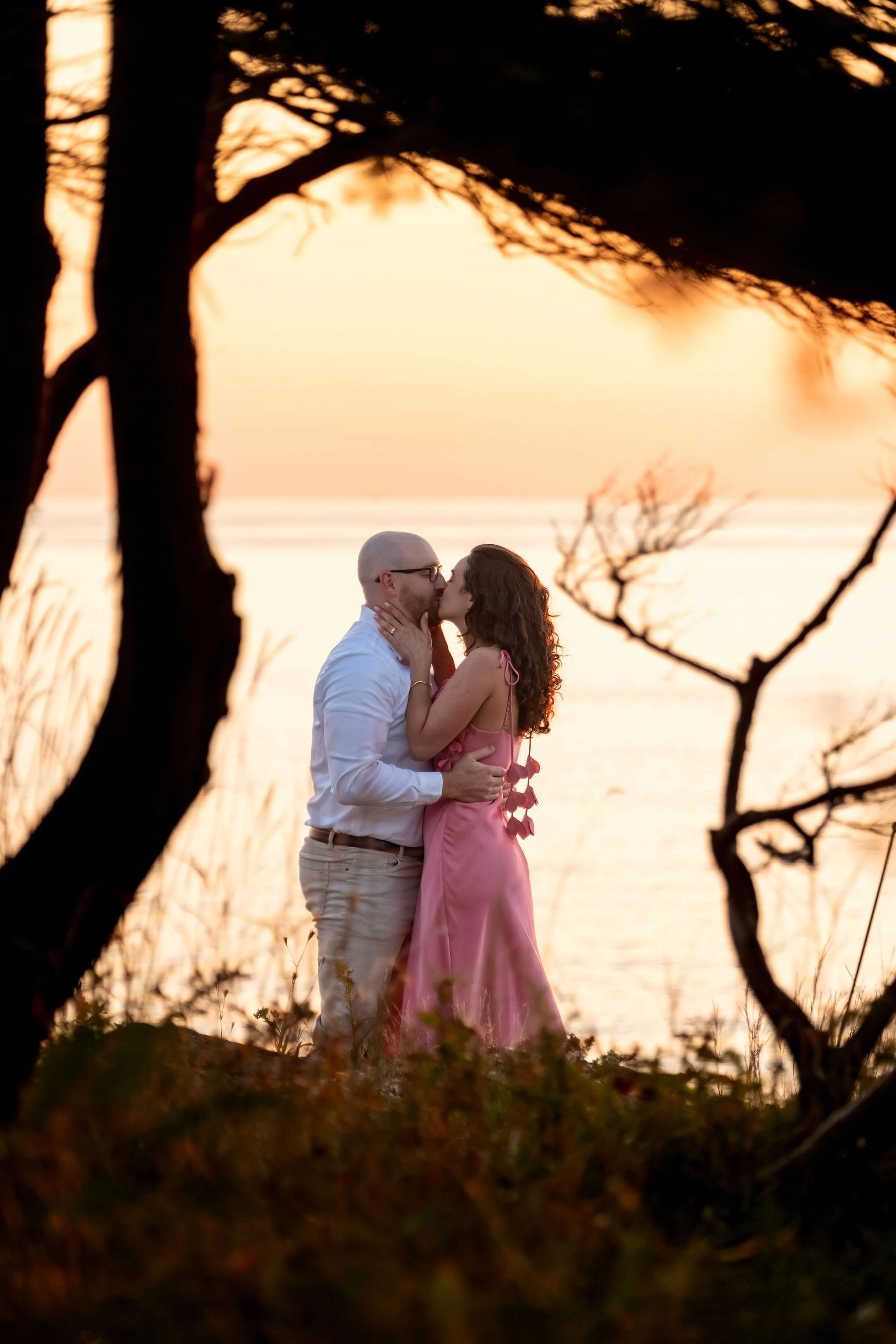Zack kissing Helena in the gap between the tree branches with the ocean glowing behind them on Marginal Way in Ogunquit Maine sunrise proposal and engagement photography VMG Productions