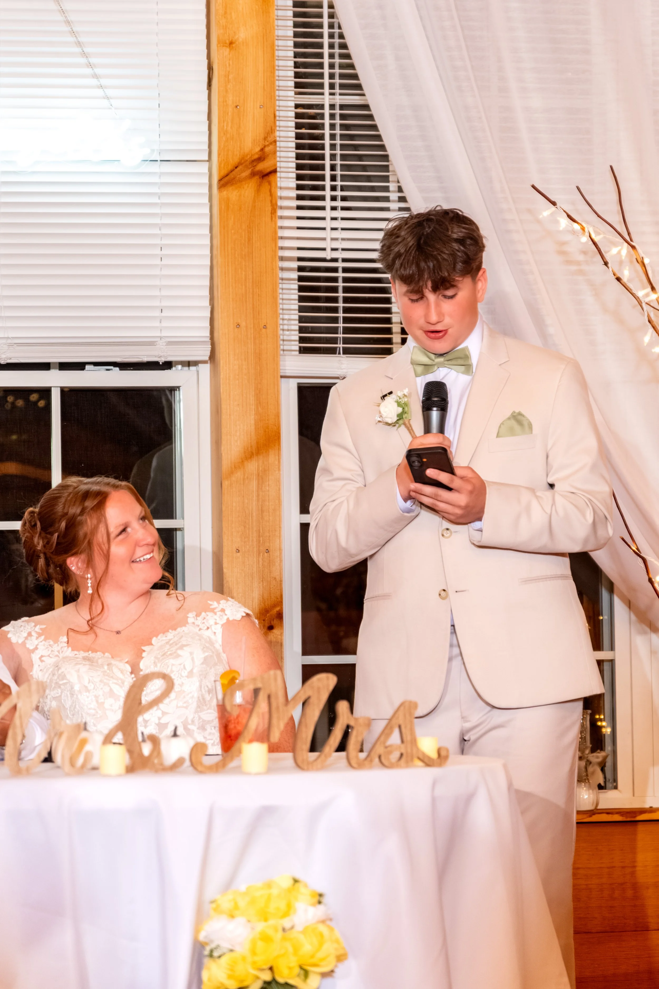 Andrew's younger brother, a best man, reading his toast off his phone as Erika smiles up at him at the Maine Wedding Barn and Event Center in Minot Maine fall wedding photography VMG Productions