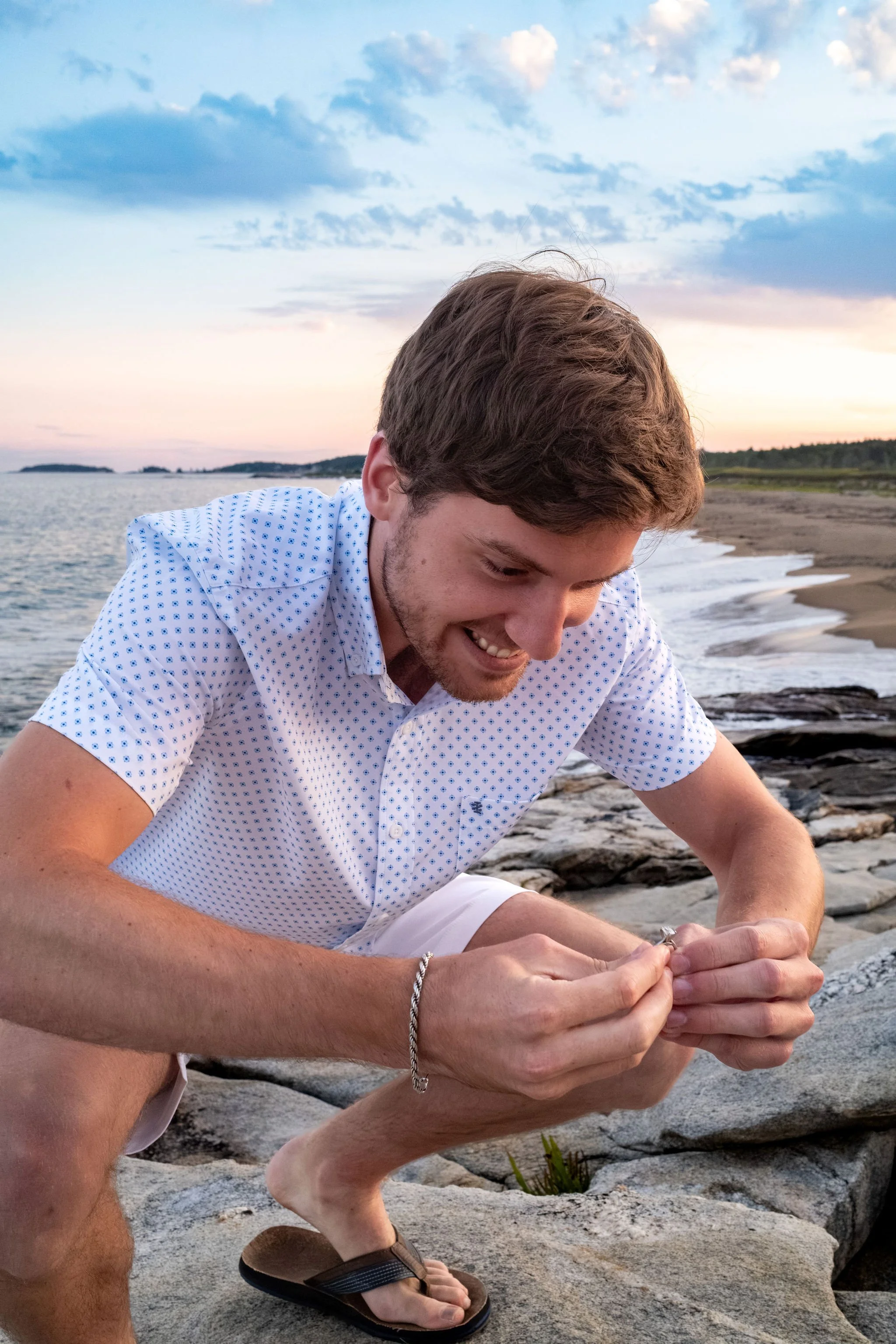 Lucas crouched on the rocks laughing with the beach behind him at Reid State Park in Georgetown Maine engagement photography VMG Productions
