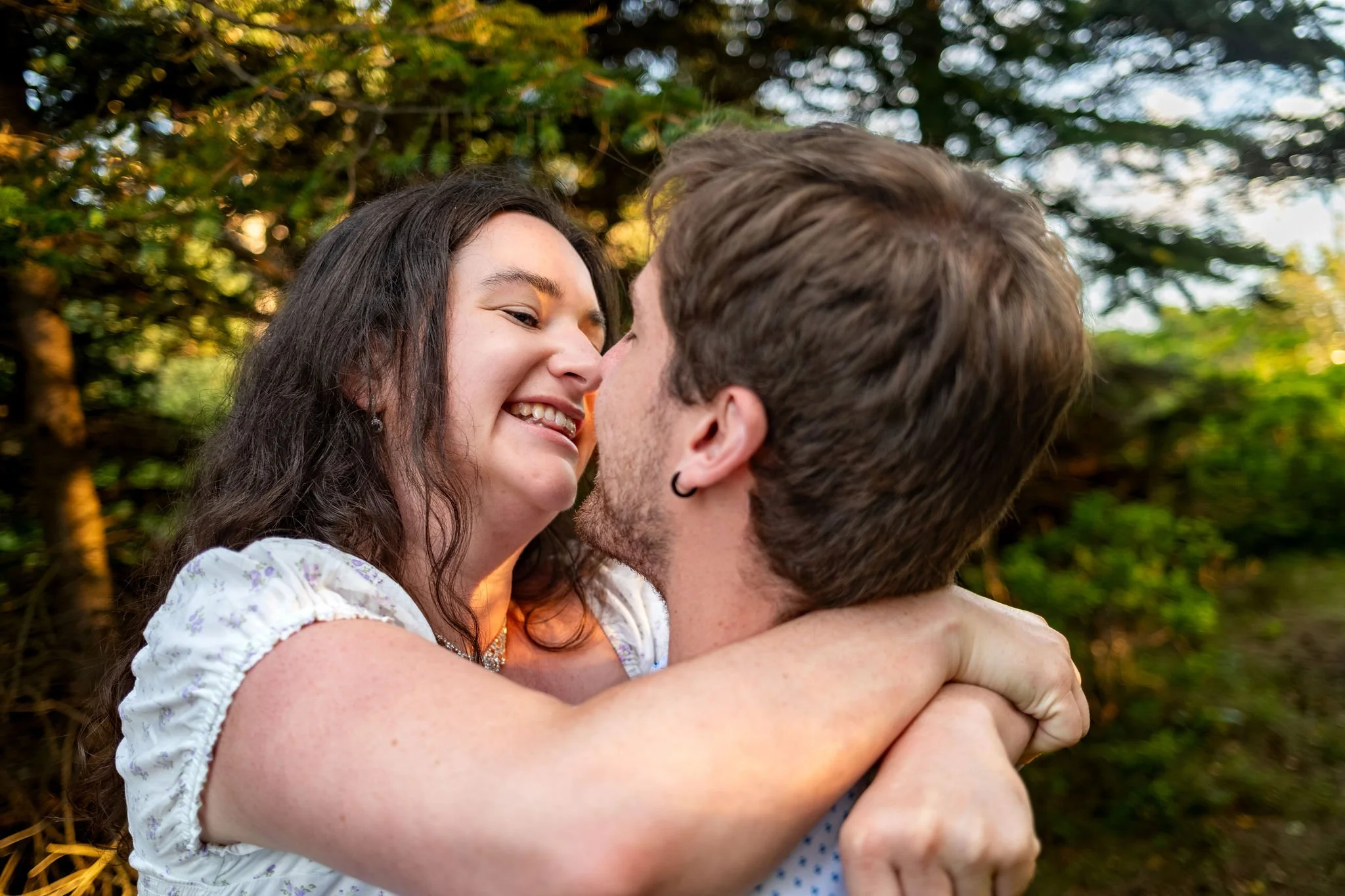 Kirstie laughing with her arms around Lucas's neck as he lifts her in the woods at Reid State Park in Georgetown Maine engagement photography VMG Productions