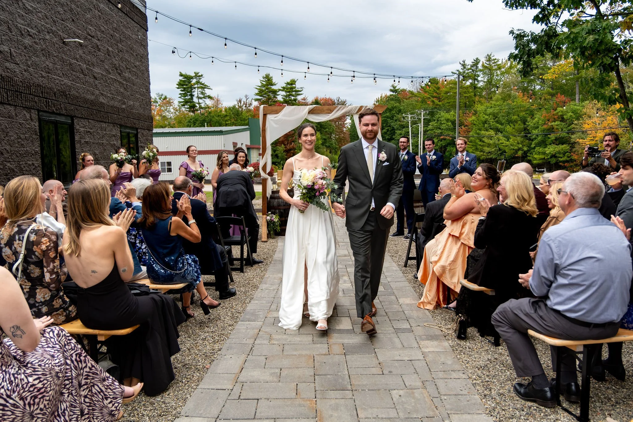 Victoria and Jack walking back up the aisle hand in hand as guests clap on both sides of the brewery courtyard at The Cellars at Allagash Brewing Company in Portland Maine fall wedding photography VMG Productions