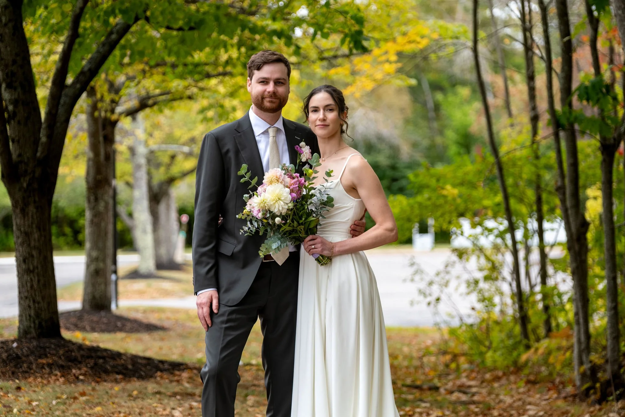 Victoria and Jack standing together under trees just starting to turn with her wildflower bouquet at The Cellars at Allagash Brewing Company in Portland Maine fall wedding photography VMG Productions
