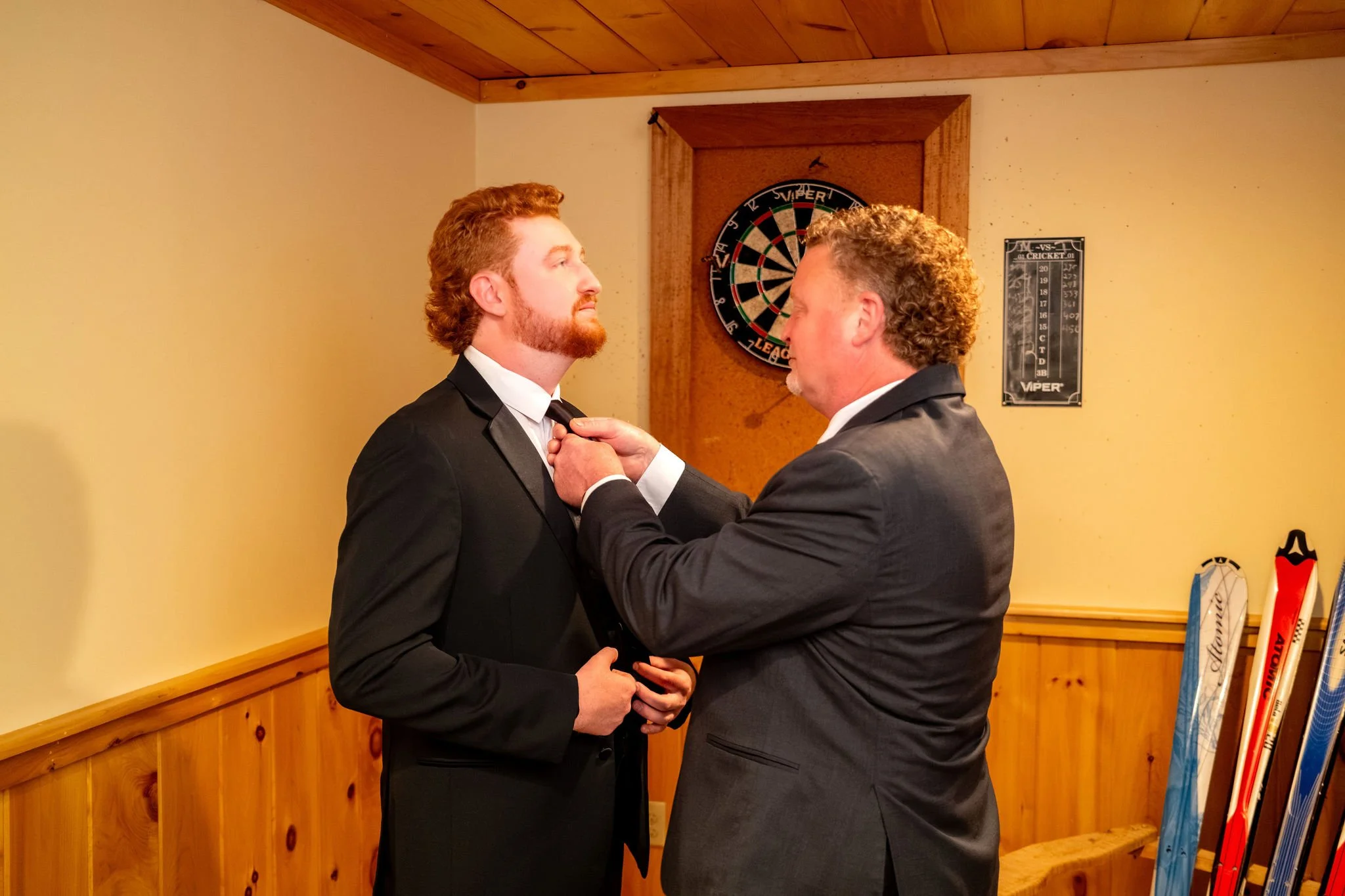 Alyssa and Colby the groom's dad straightening his tie in front of the basement dartboard in Bethel Maine summer wedding photography VMG Productions