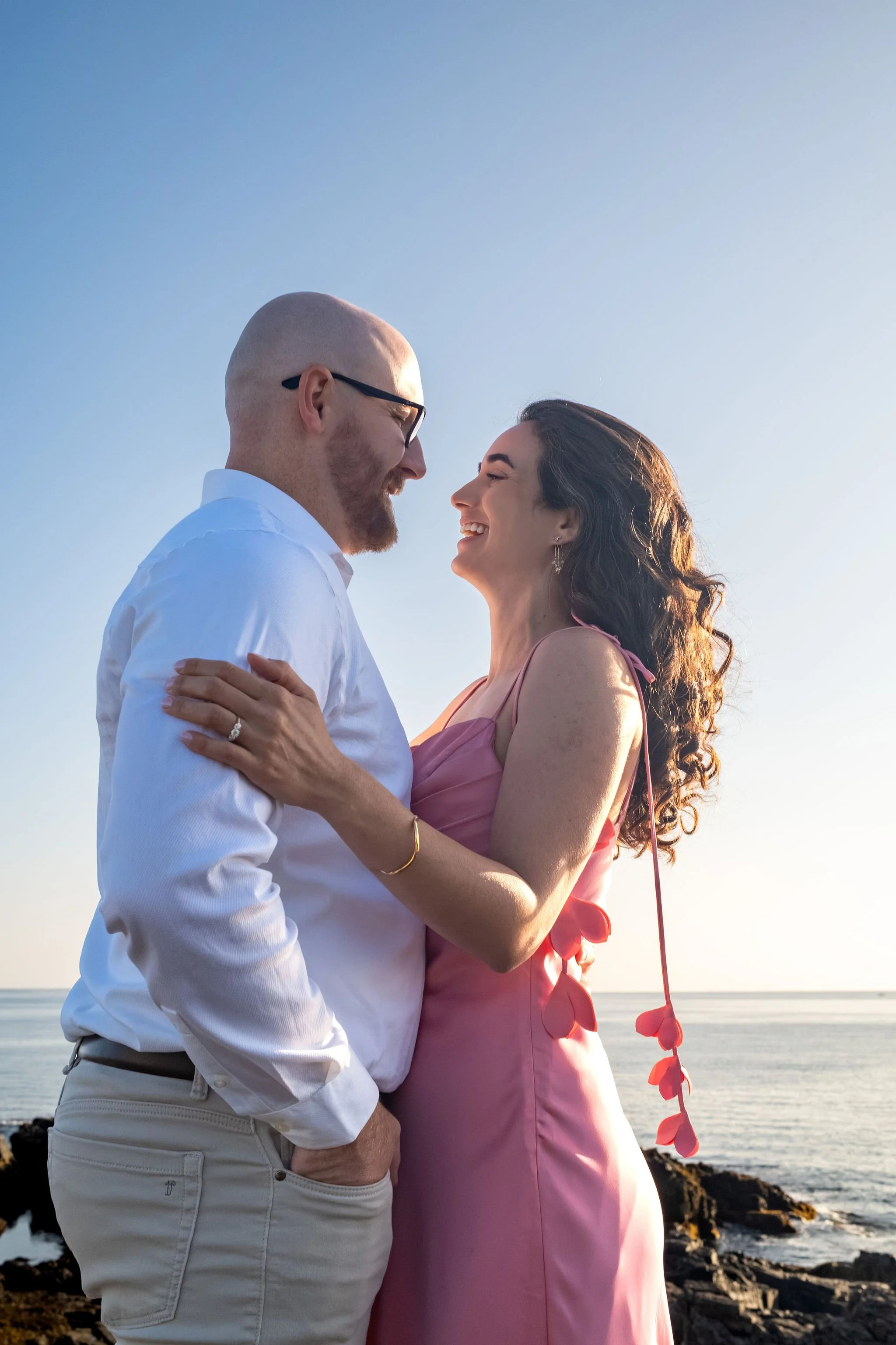 Helena laughing up at Zack as they stand close with the ocean behind them on Marginal Way in Ogunquit Maine sunrise proposal and engagement photography VMG Productions