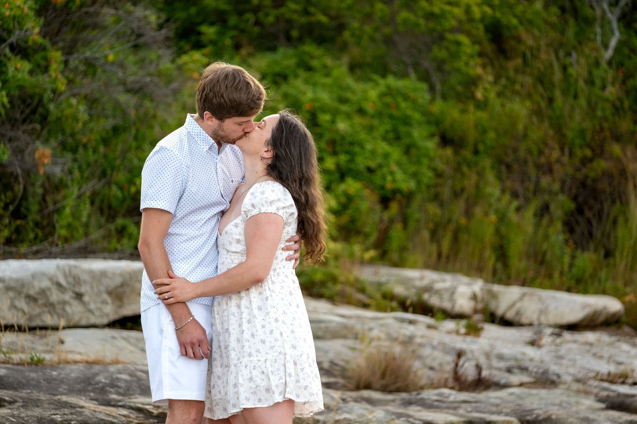 Lucas holding Kirstie close for a kiss in front of the shoreline greenery at Reid State Park in Georgetown Maine engagement photography VMG Productions