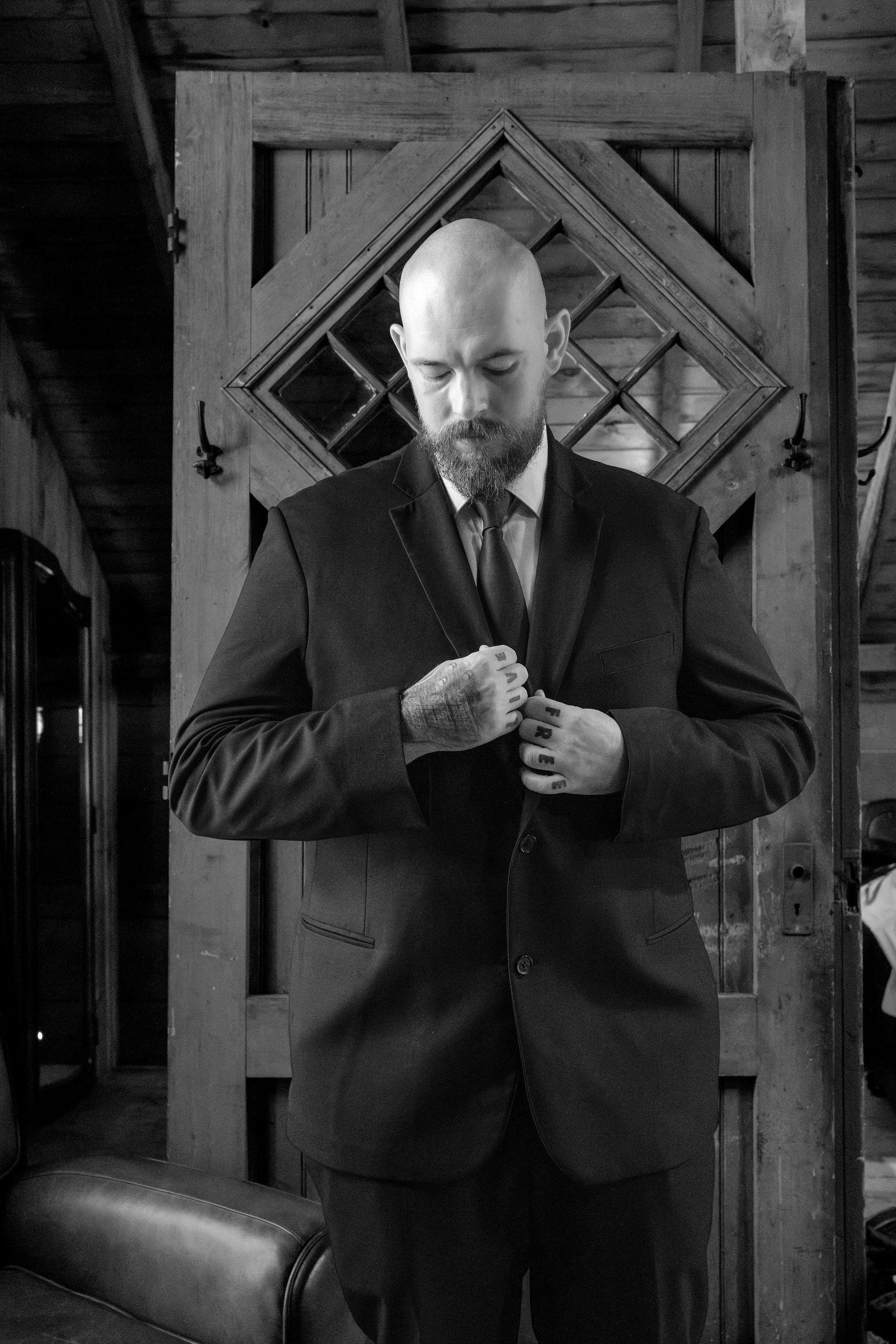 Black and white portrait of Sam looking down as he buttons his suit jacket in the barn at Boothbay Playhouse in Boothbay Maine fall wedding photography VMG Productions