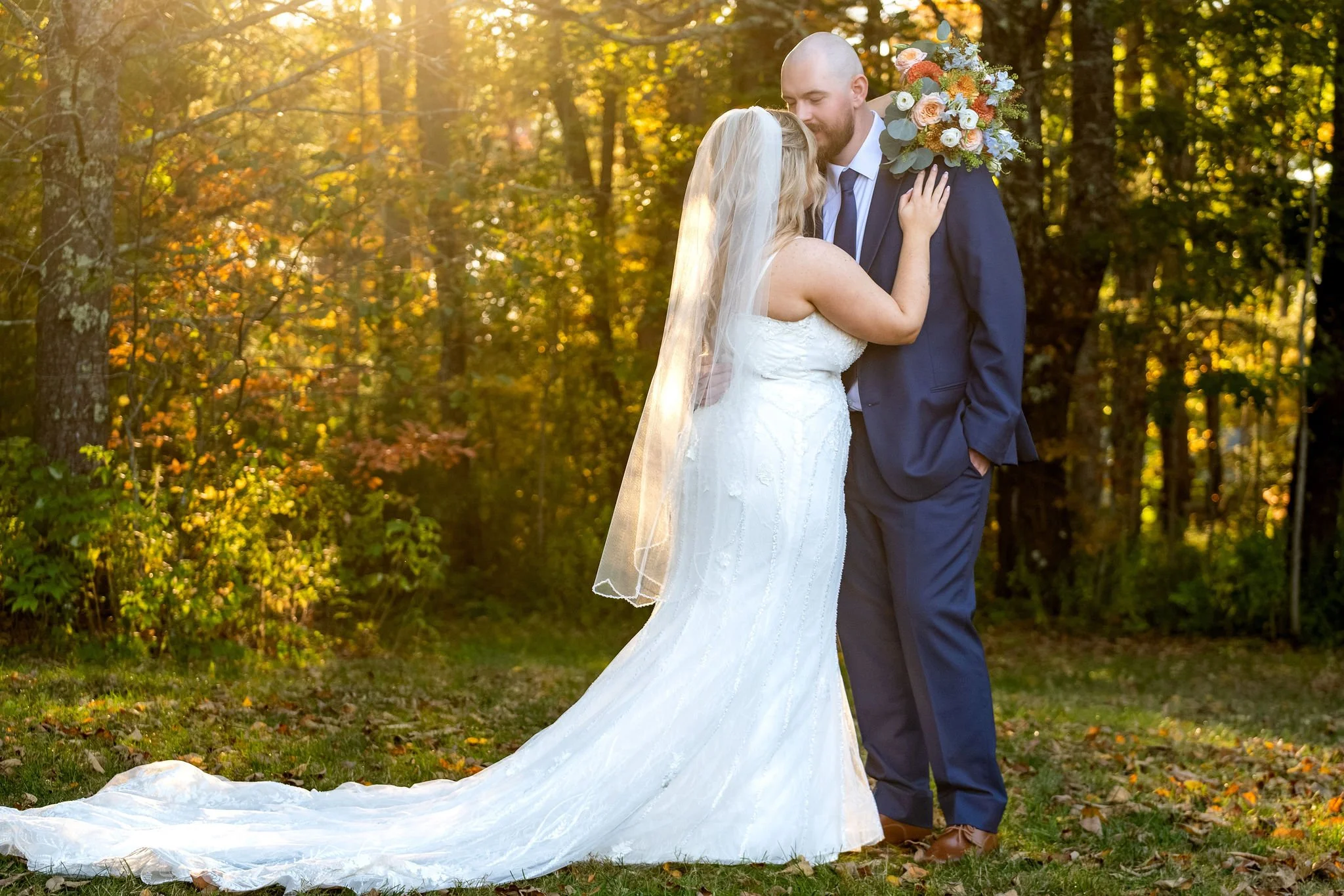 Ellie and Sam embracing as the golden sun backlights the trees and her veil at Boothbay Playhouse in Boothbay Maine fall wedding photography VMG Productions