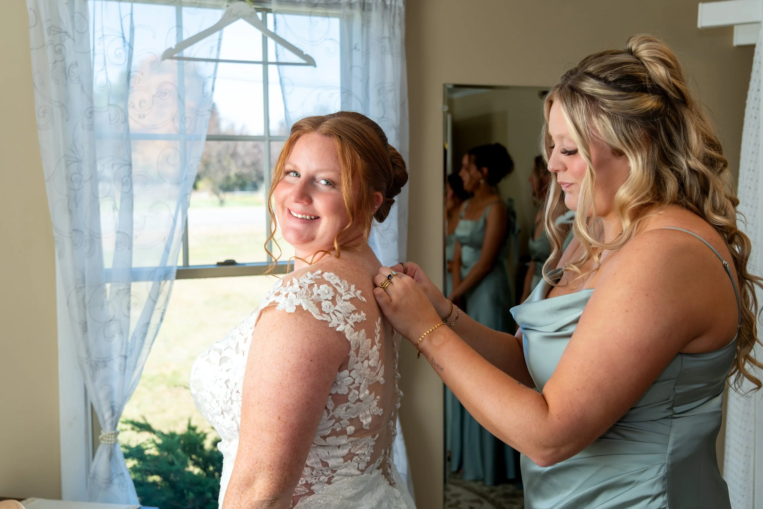 A bridesmaid helping Erika into her lace wedding dress by the window at the Maine Wedding Barn and Event Center in Minot Maine fall wedding photography VMG Productions